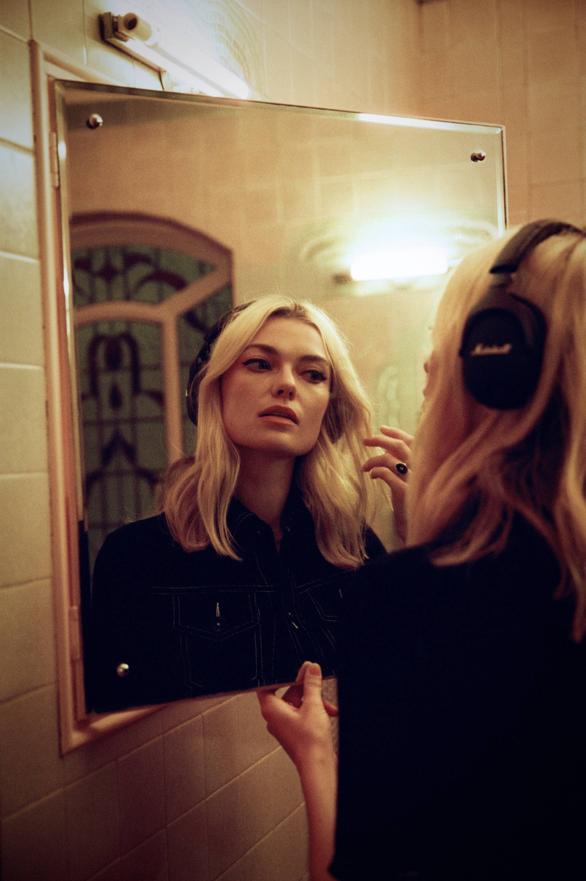 Eine Frau mit blonden Haaren trägt Kopfhörer und schaut in einen Spiegel in einem warm beleuchteten Raum.