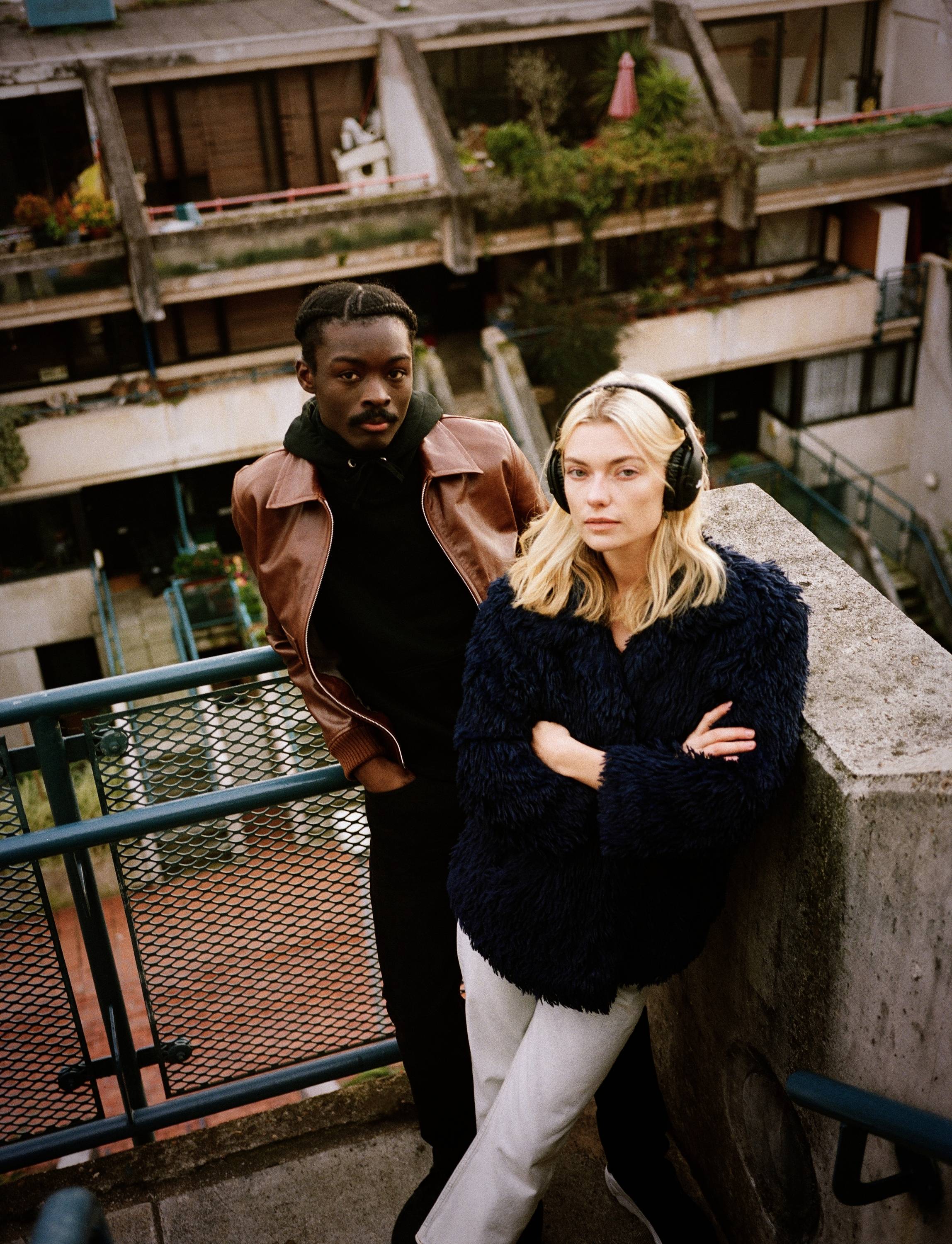 Zwei Personen stehen auf einem Balkon. Eine Person trägt Kopfhörer und eine blaue Jacke, die andere eine braune Lederjacke. Urbaner Hintergrund.