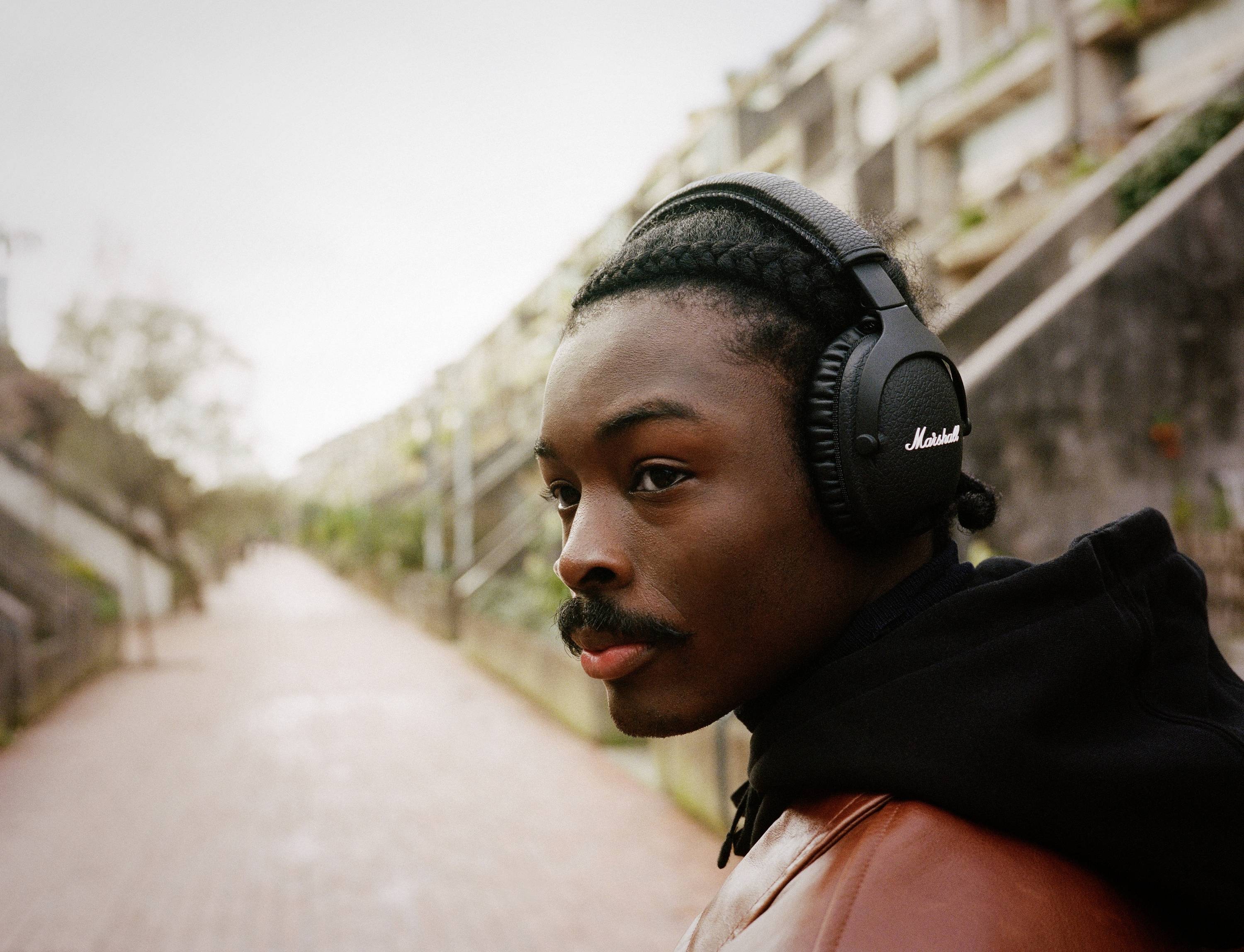 Eine Person mit Kopfhörern spaziert auf einem gepflasterten Weg entlang. Im Hintergrund sind Bäume und Gebäude zu sehen.