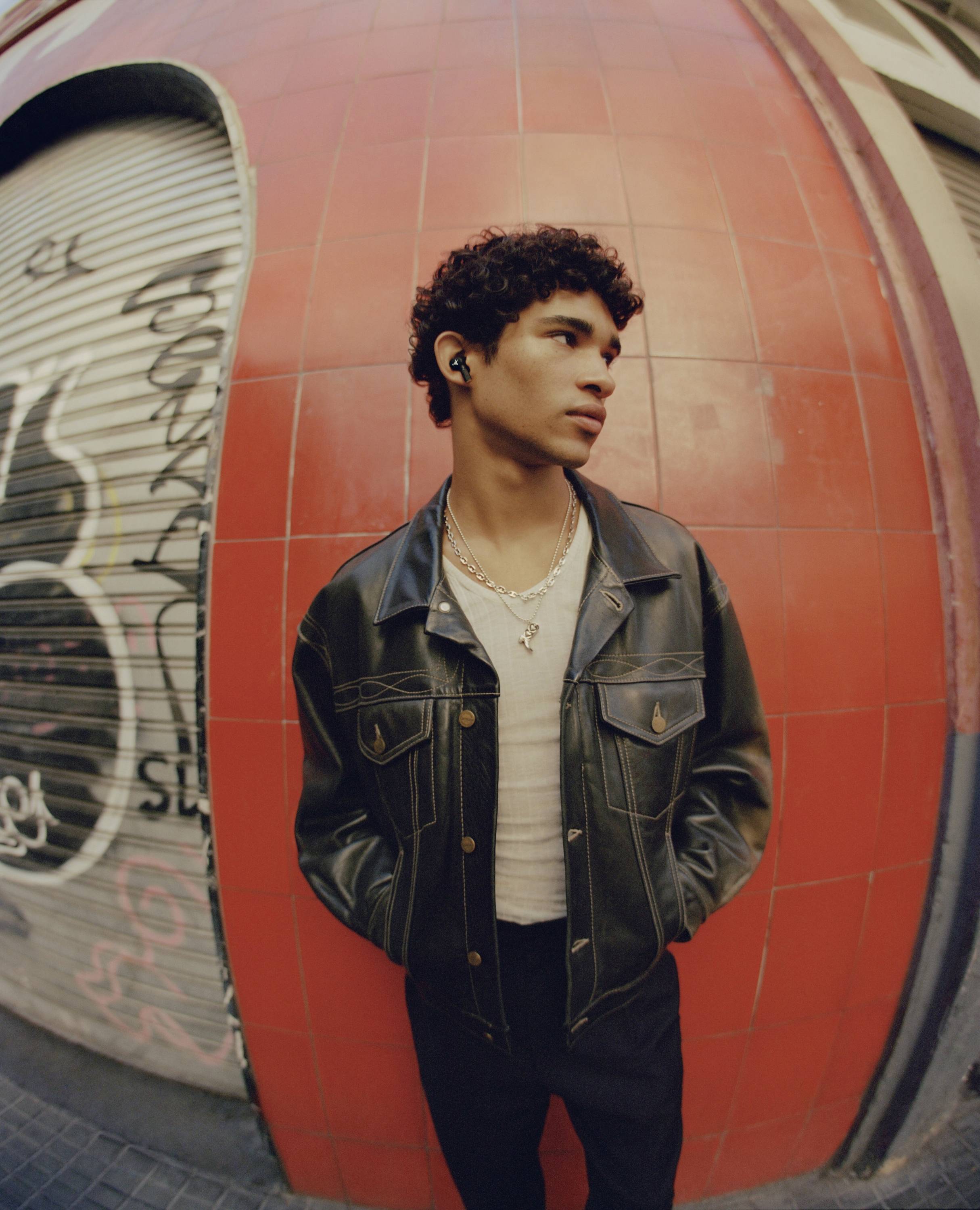 Eine Person mit schwarzer Jacke steht vor einer großen roten Fliese und grauer, mit Graffiti besprühter Wand. Sie hat kurzes, lockiges Haar.