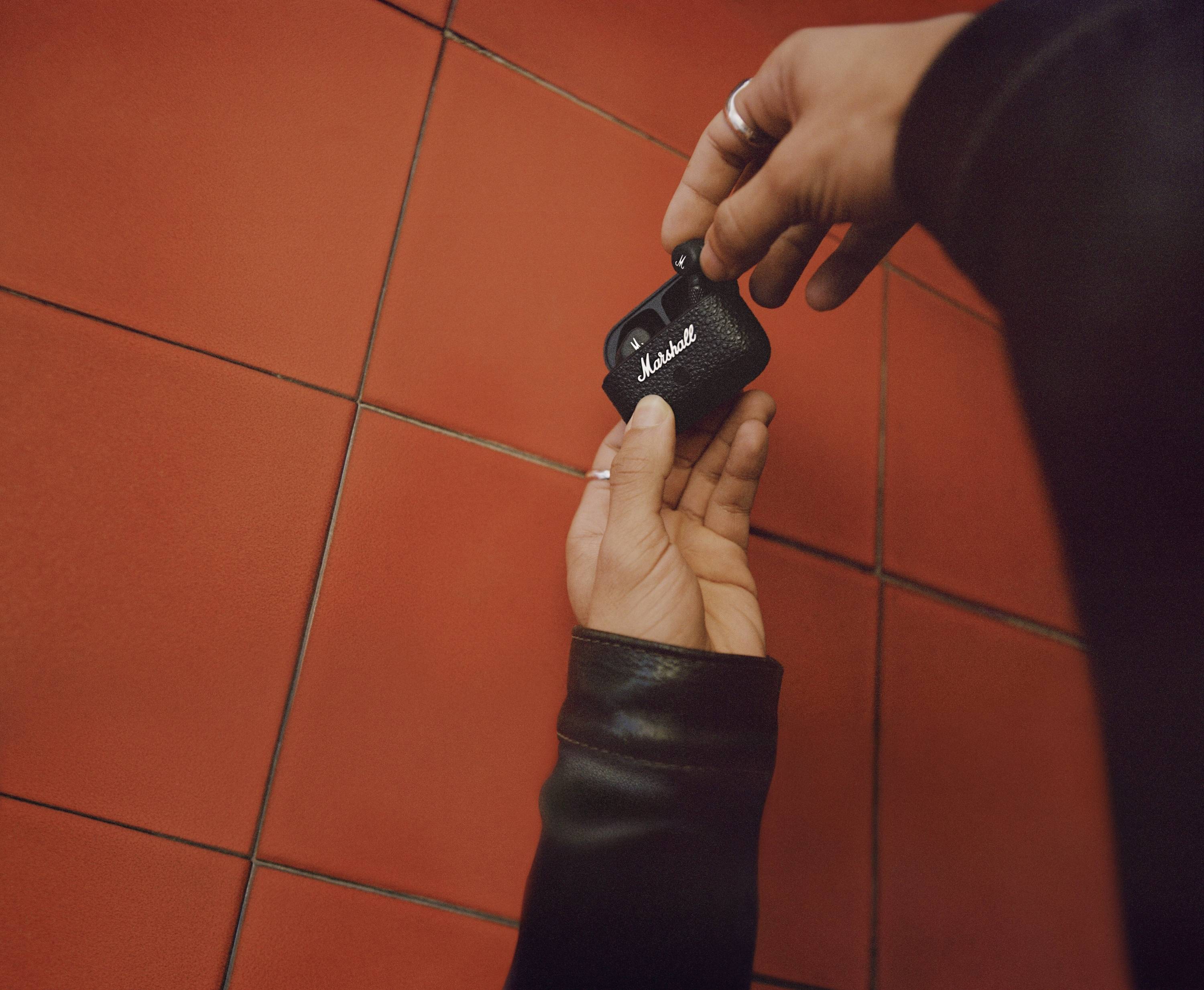 Eine Person in schwarzer Kleidung hält ein kleines, schwarzes Marshall-Gerät vor einem roten Fliesenhintergrund.