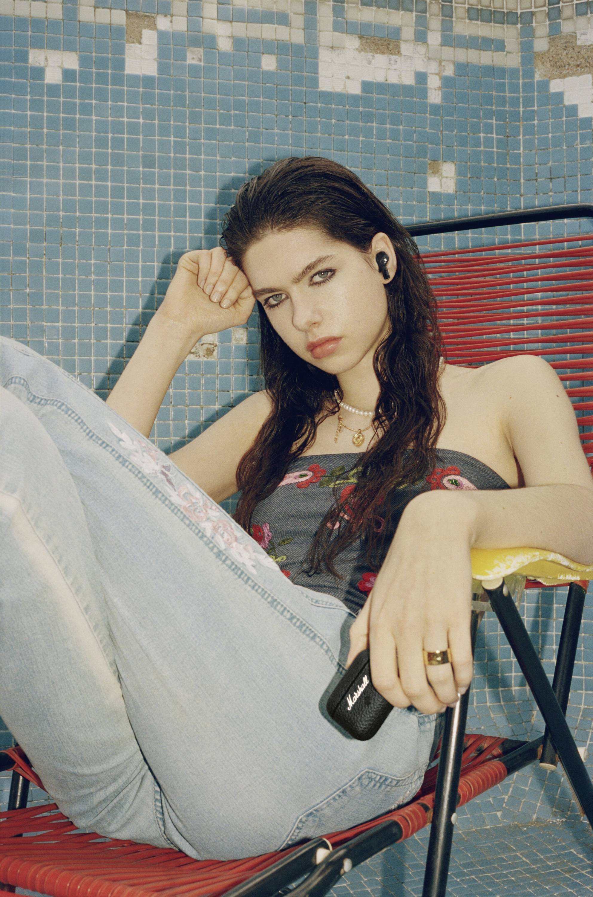 Junge Frau sitzt lässig auf einem roten Stuhl vor blauer gefliester Wand, hält ein kleines schwarzes Gerät. Sie trägt Kopfhörer.