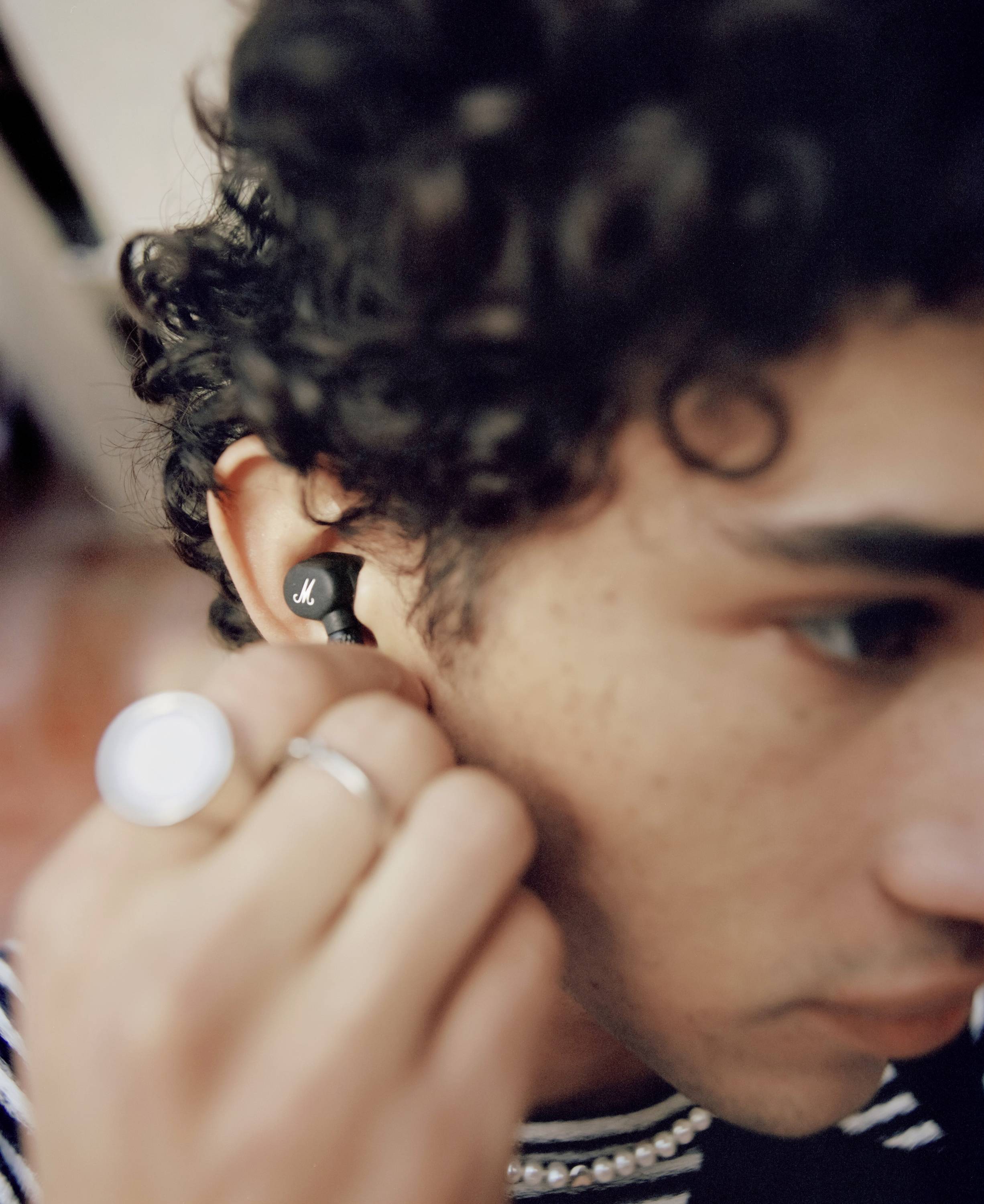 Eine Person mit lockigem Haar trägt kabellose Ohrhörer und hält eine Hand daran. Der Fokus liegt auf dem Ohr und den Ohrhörern.