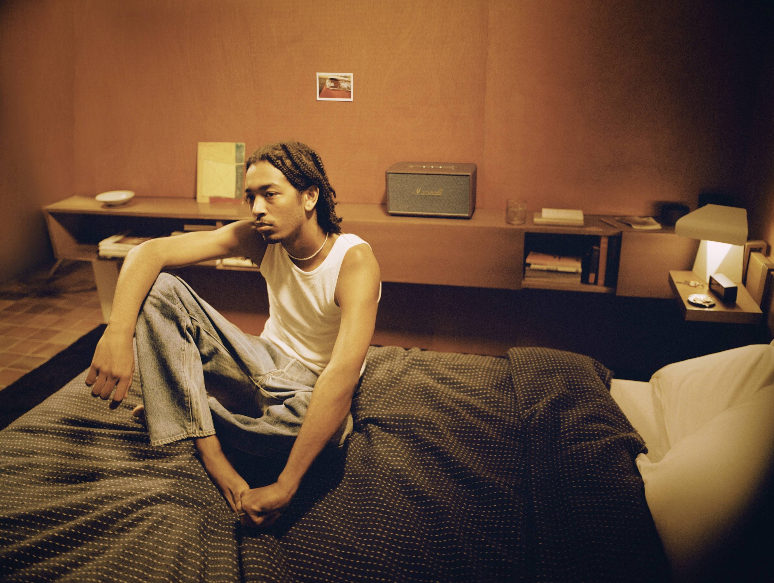 Eine Person sitzt nachdenklich auf einem Bett in einem gemütlichen, schummrig beleuchteten Zimmer mit modernen Regalen und Radio.