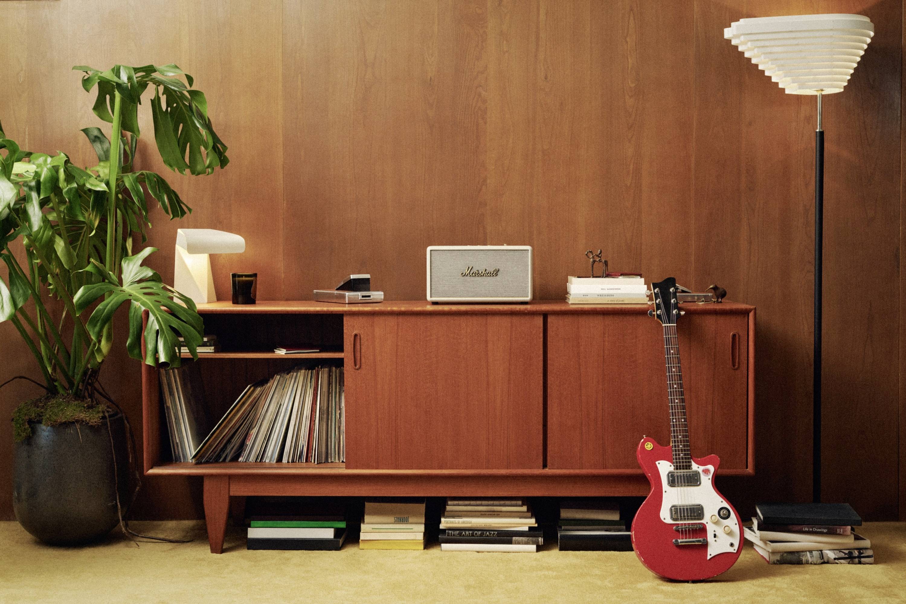 Ein Wohnzimmer mit einem hölzernen Sideboard. Darauf steht ein Lautsprecher, Schallplatten sind offen. Rechts lehnt eine rote E-Gitarre. Links steht eine Topfpflanze.
