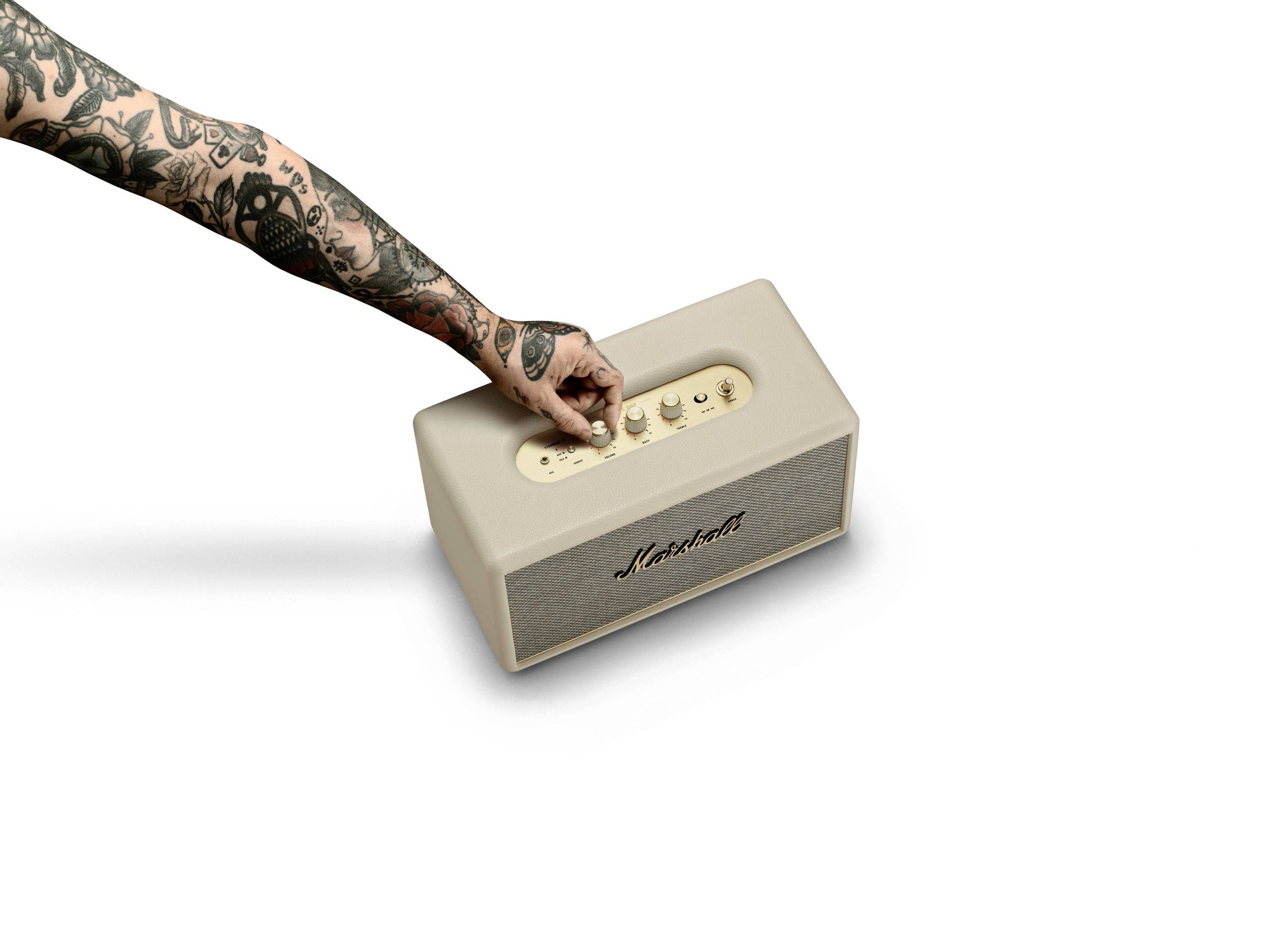 Ein tätowierter Arm stellt die Lautstärke an einem beigen Lautsprecher mit der Aufschrift 'Marshall' ein.