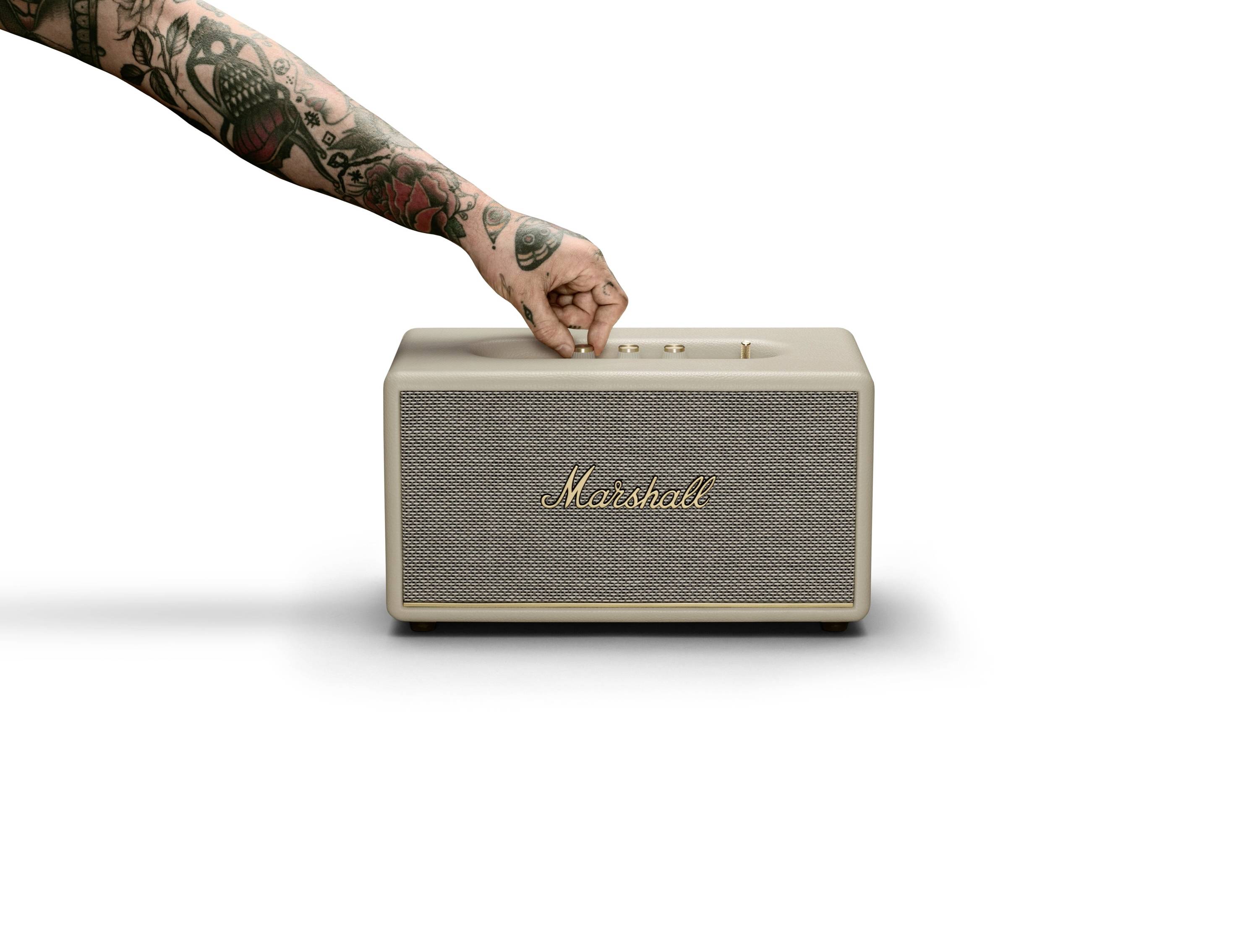 Eine tätowierte Hand greift nach einem tragbaren Lautsprecher mit der Aufschrift 'Marshall' auf einem weißen Hintergrund.