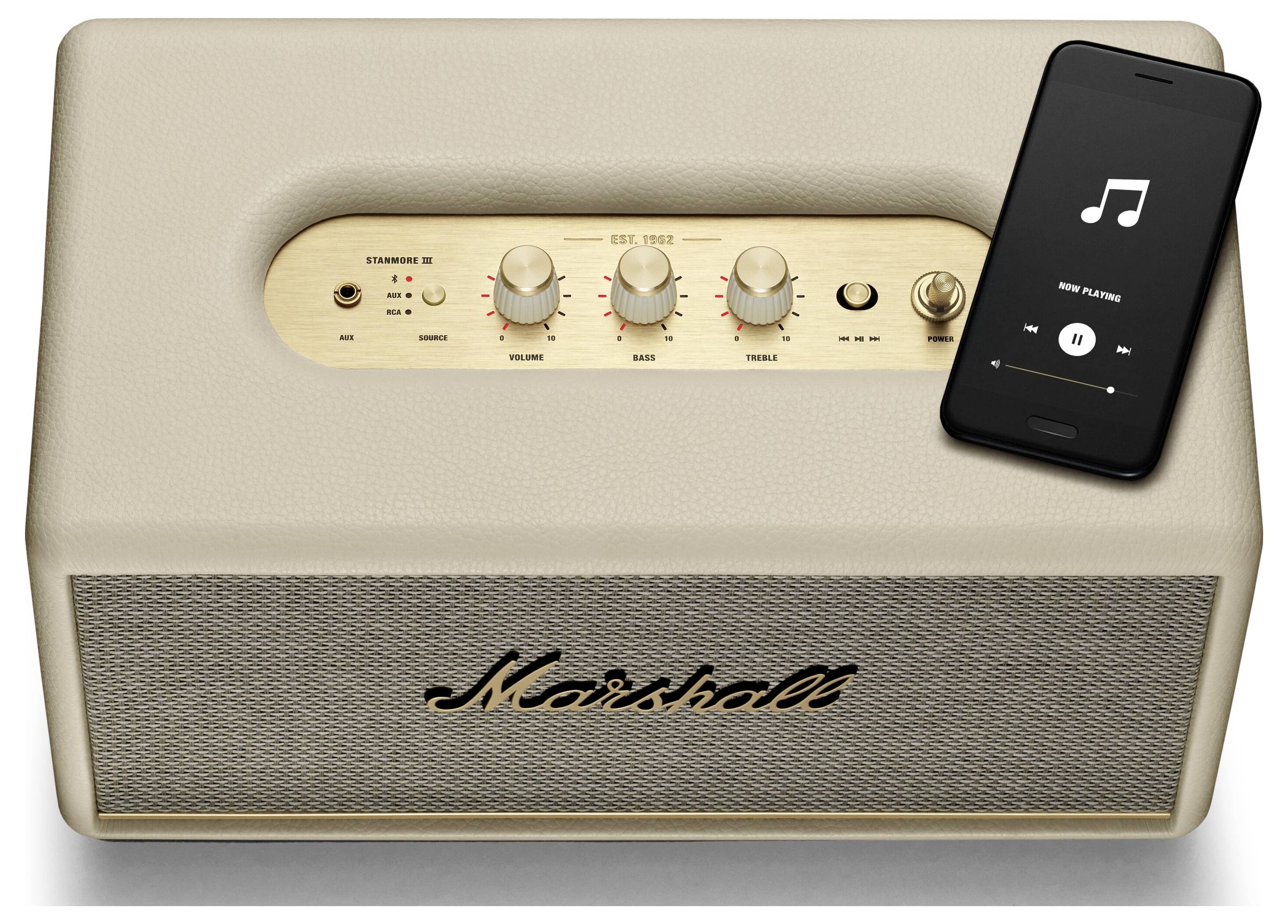 Ein cremefarbener Lautsprecher mit goldenem Bedienfeld, Lautstärkeregler in der Mitte. Oben liegt ein Smartphone mit geöffneter Musik-App.
