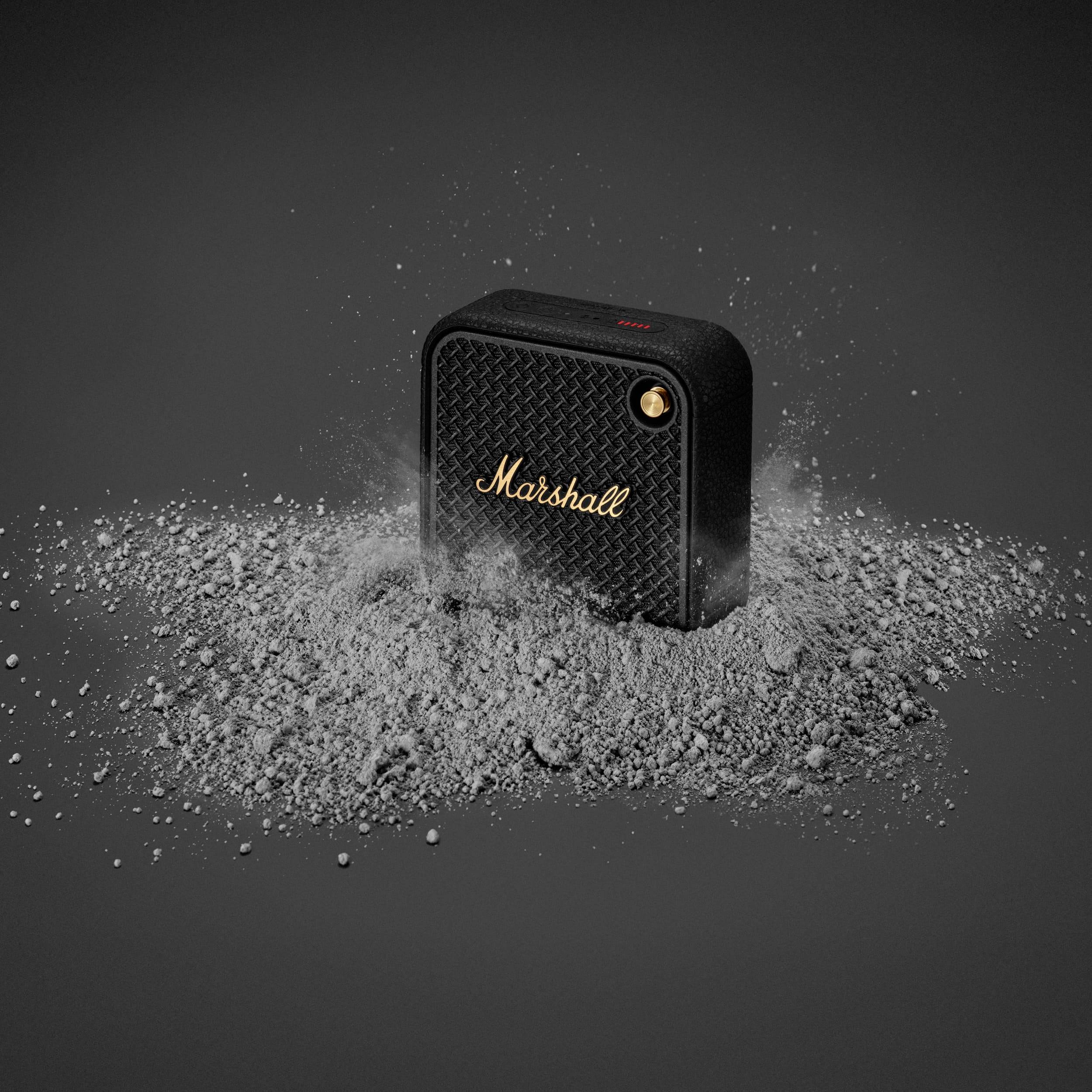 'Schwarzer Marshall-Lautsprecher, umgeben von aufgewirbeltem Staub. Kompakt und robust, mit goldfarbenem Logo auf der Vorderseite.'