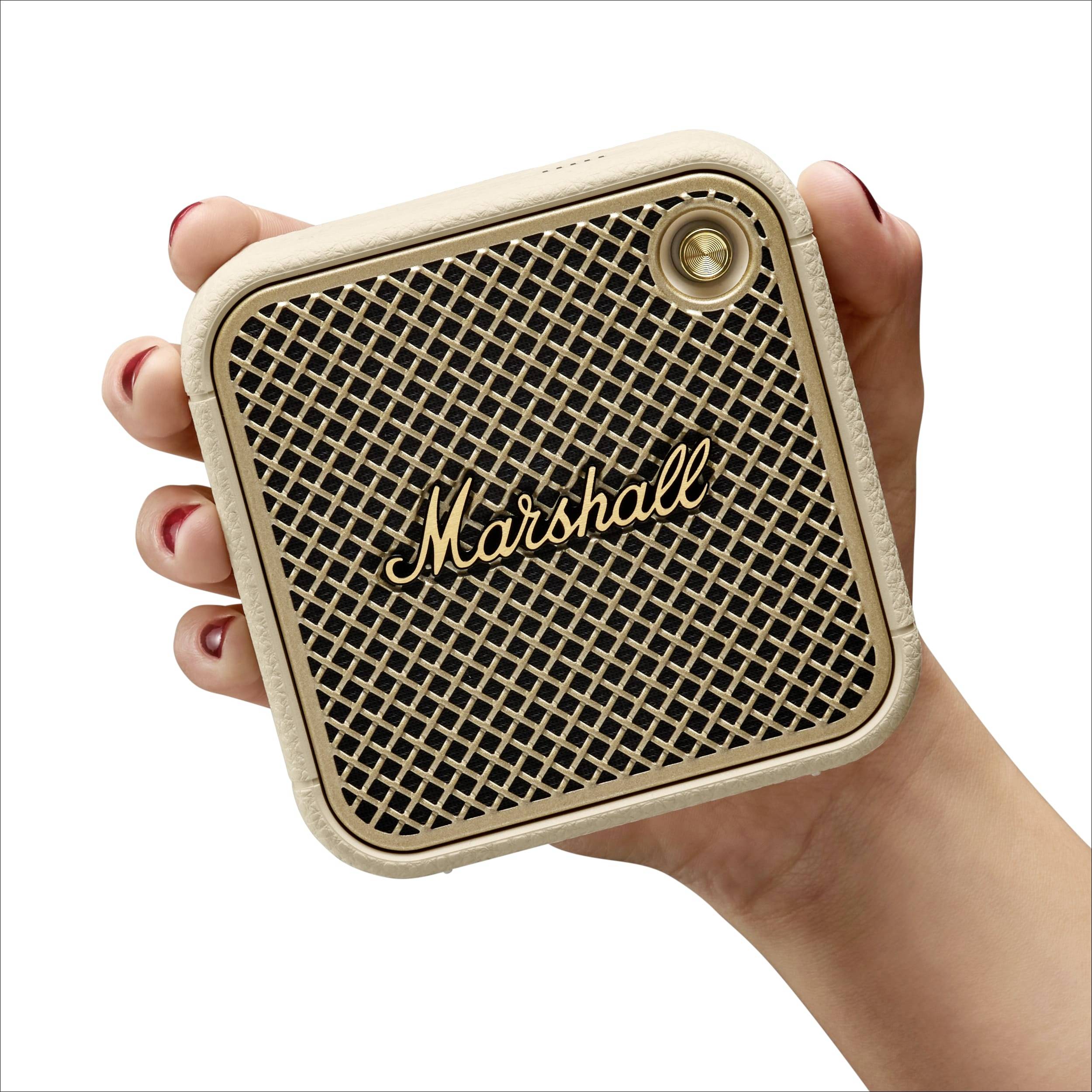 Eine Hand hält einen kompakten, quadratischen Lautsprecher mit einem goldenen 'Marshall'-Logo und einem Gittermuster auf der Vorderseite.