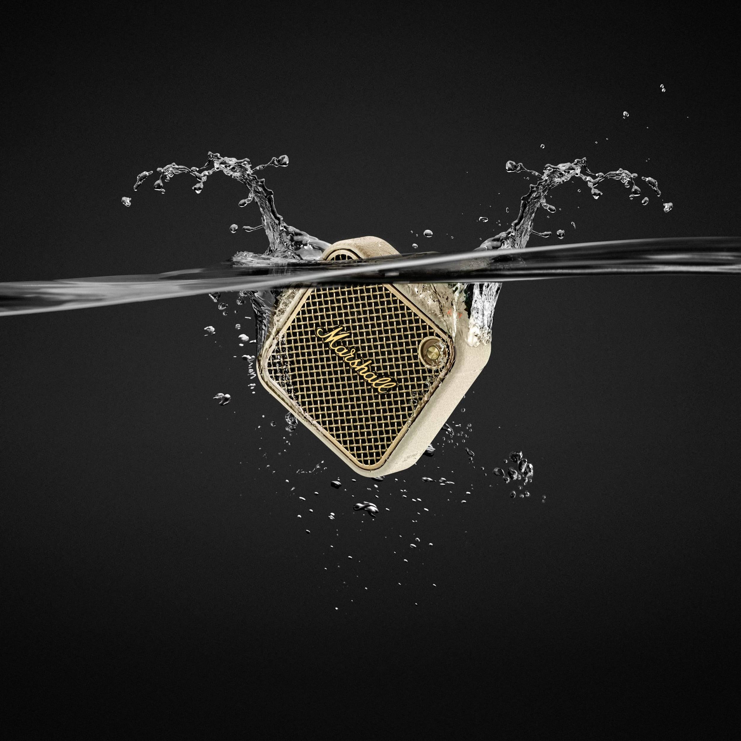 Ein tragbarer Lautsprecher wird in Wasser getaucht, umgeben von spritzenden Tropfen. Das Gehäuse ist beige und hat ein Metallgitter.