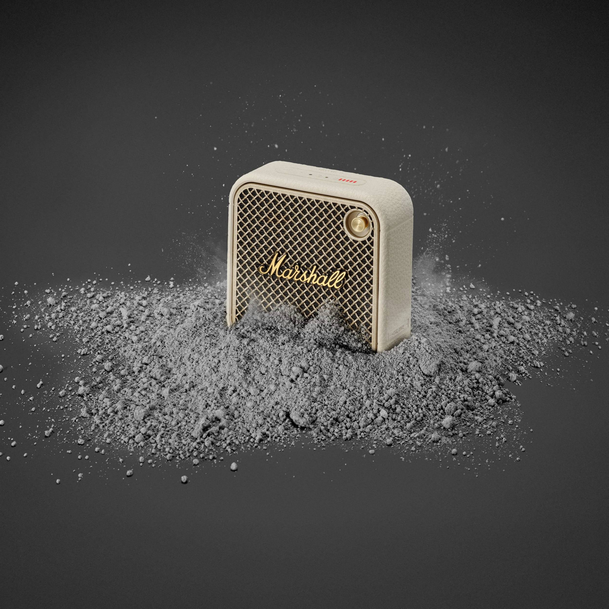 Ein beiger Bluetooth-Lautsprecher mit der Aufschrift 'Marshall' steht vor einem dunklen Hintergrund, umgeben von grauem Staub.