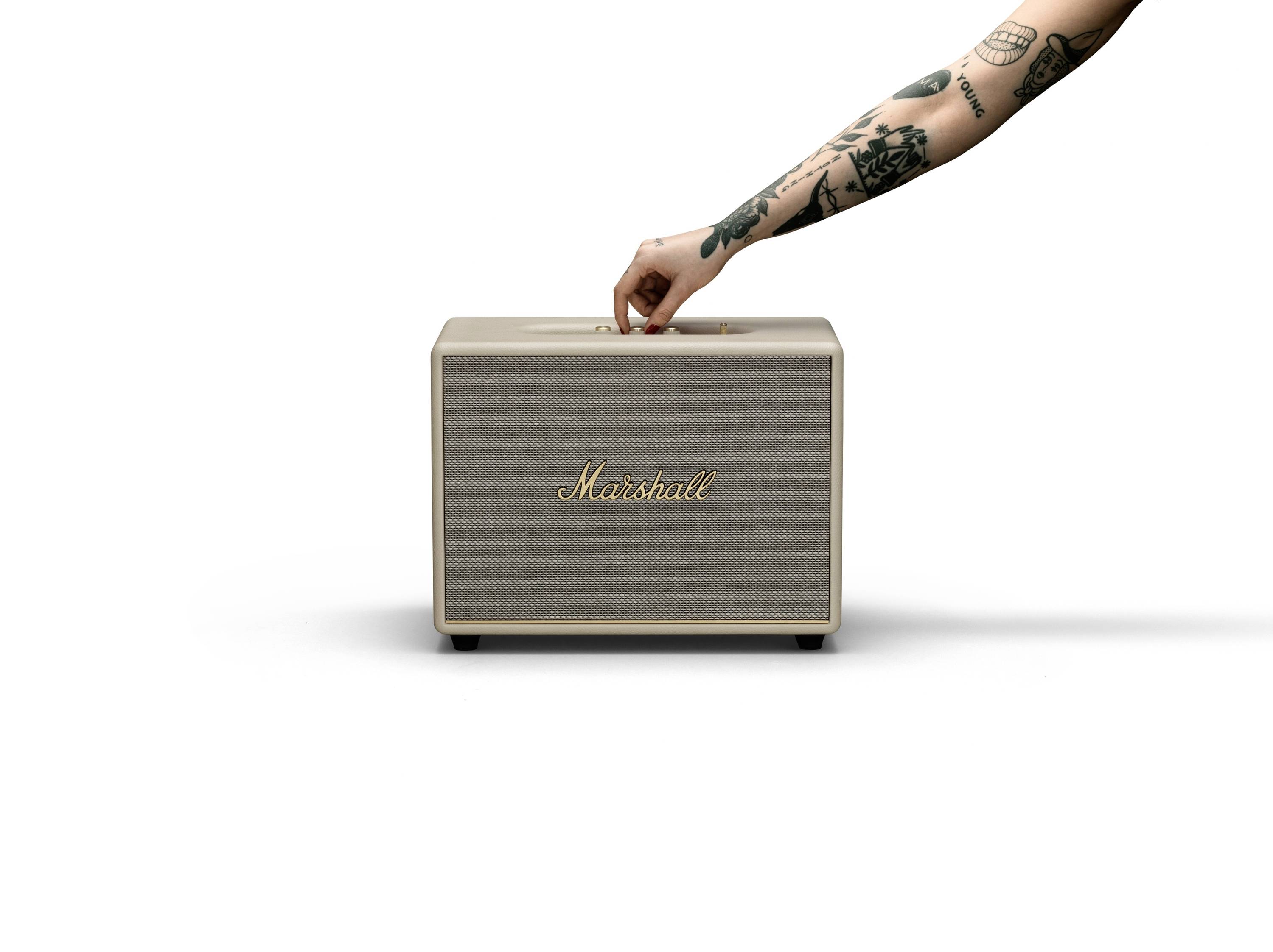 Ein tätowierter Arm bedient einen hellbraunen Lautsprecher mit der Aufschrift 'Marshall' auf weißem Hintergrund.