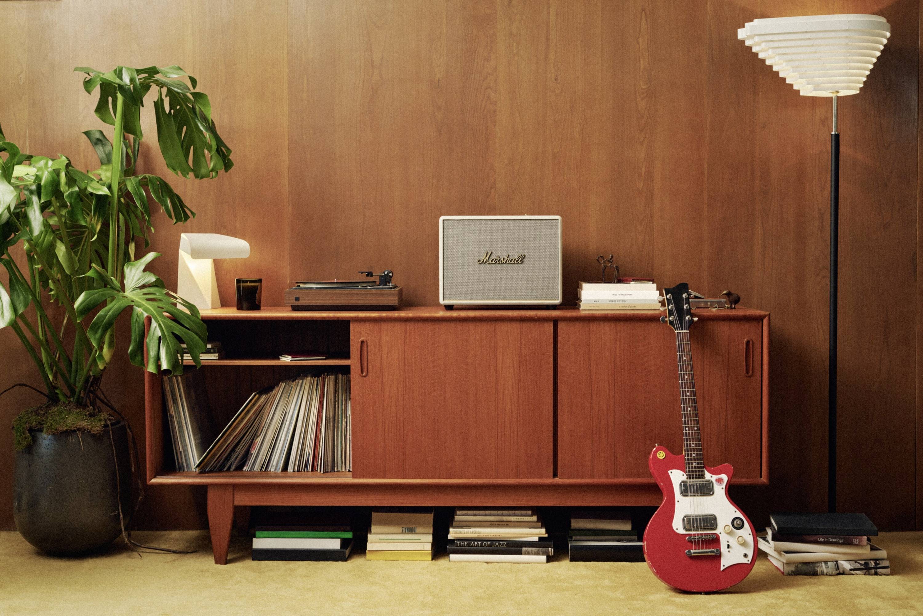 Eine rote E-Gitarre steht vor einem Vintage-Holzregal mit einem Plattenspieler, einem Lautsprecher und Platten. Daneben eine Lampe und eine Pflanze.