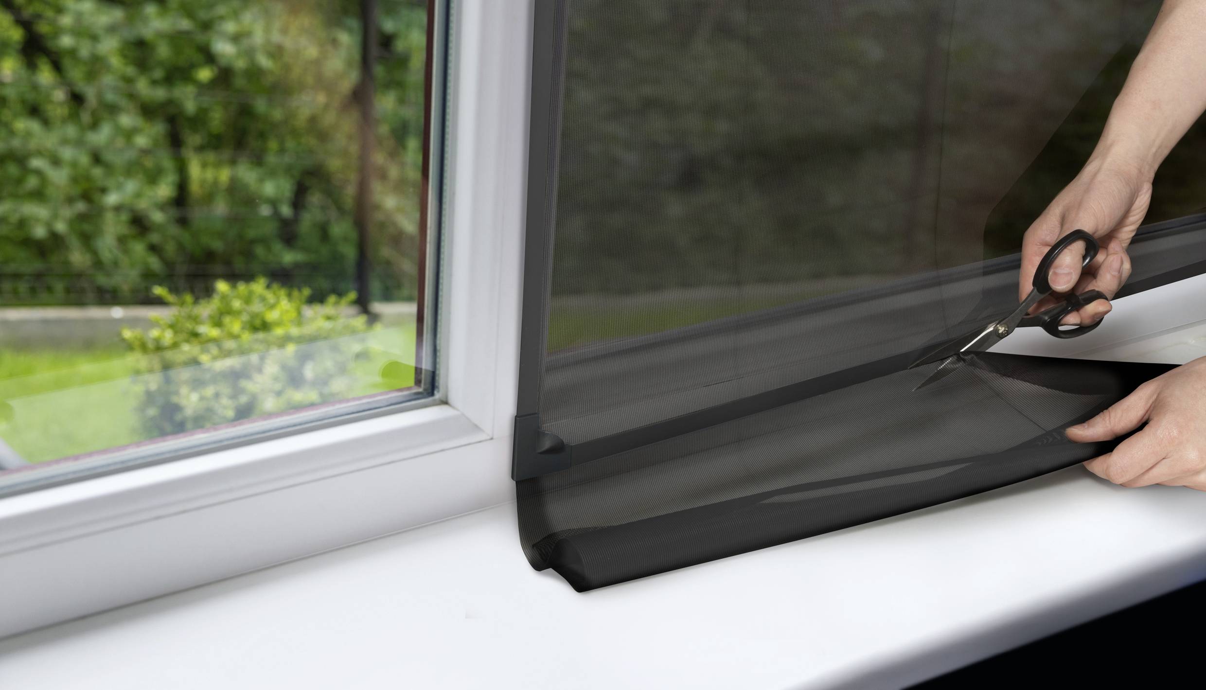 Person schneidet schwarze Fensterfolie auf Fensterbank. Draußen ist ein grüner Garten zu sehen.