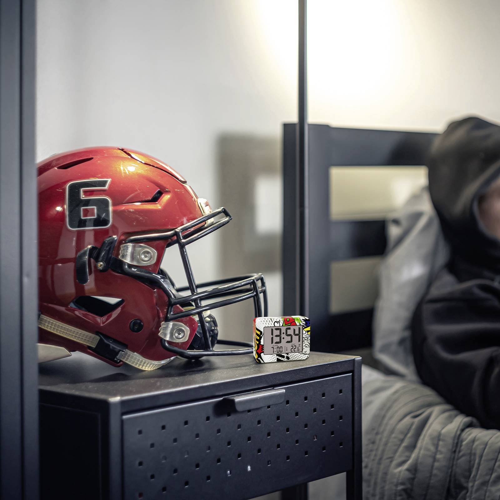 Ein roter American-Football-Helm mit der Nummer 6 liegt auf einem schwarzen Metalltisch neben einem digitalen Wecker, der 13:54 anzeigt.