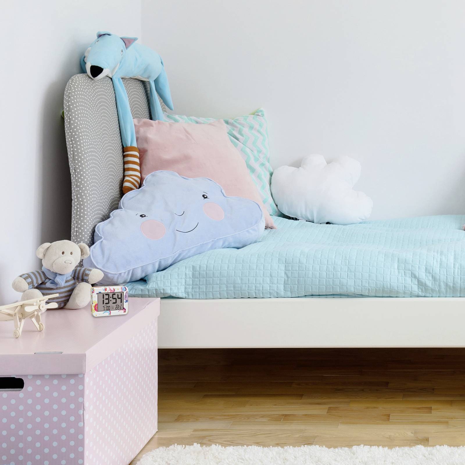 Kinderzimmer mit einem Bett, das mit farbenfrohen Kissen in Wolken- und Sternform dekoriert ist. Ein Stofftier hängt am Kopfende. Links eine rosa gepunktete Spielzeugkiste.