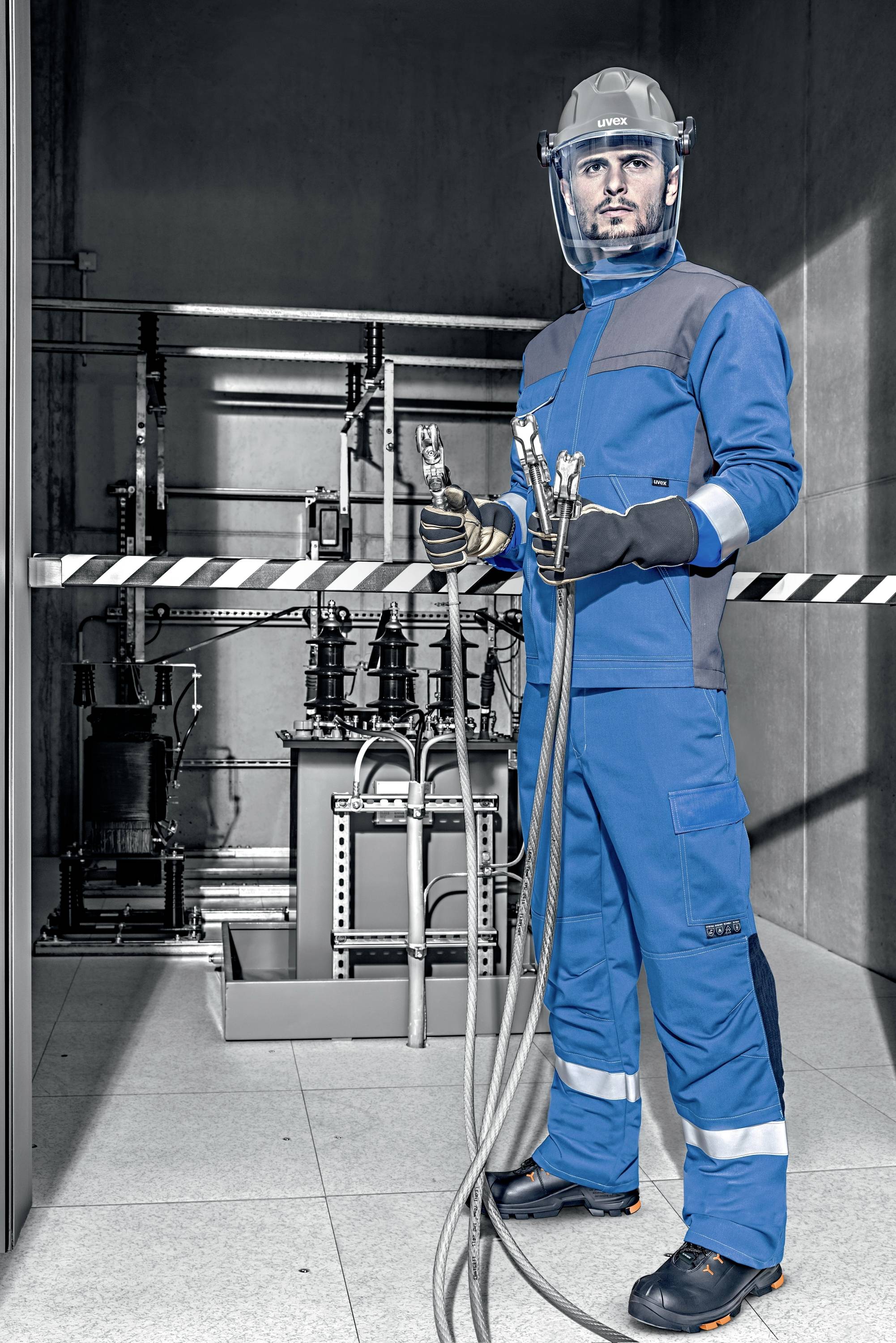 Ein Mann in blauer Schutzkleidung hält Kabel und steht vor industriellen Maschinen. Er trägt Helm und Schutzvisier in einer grauen Umgebung.