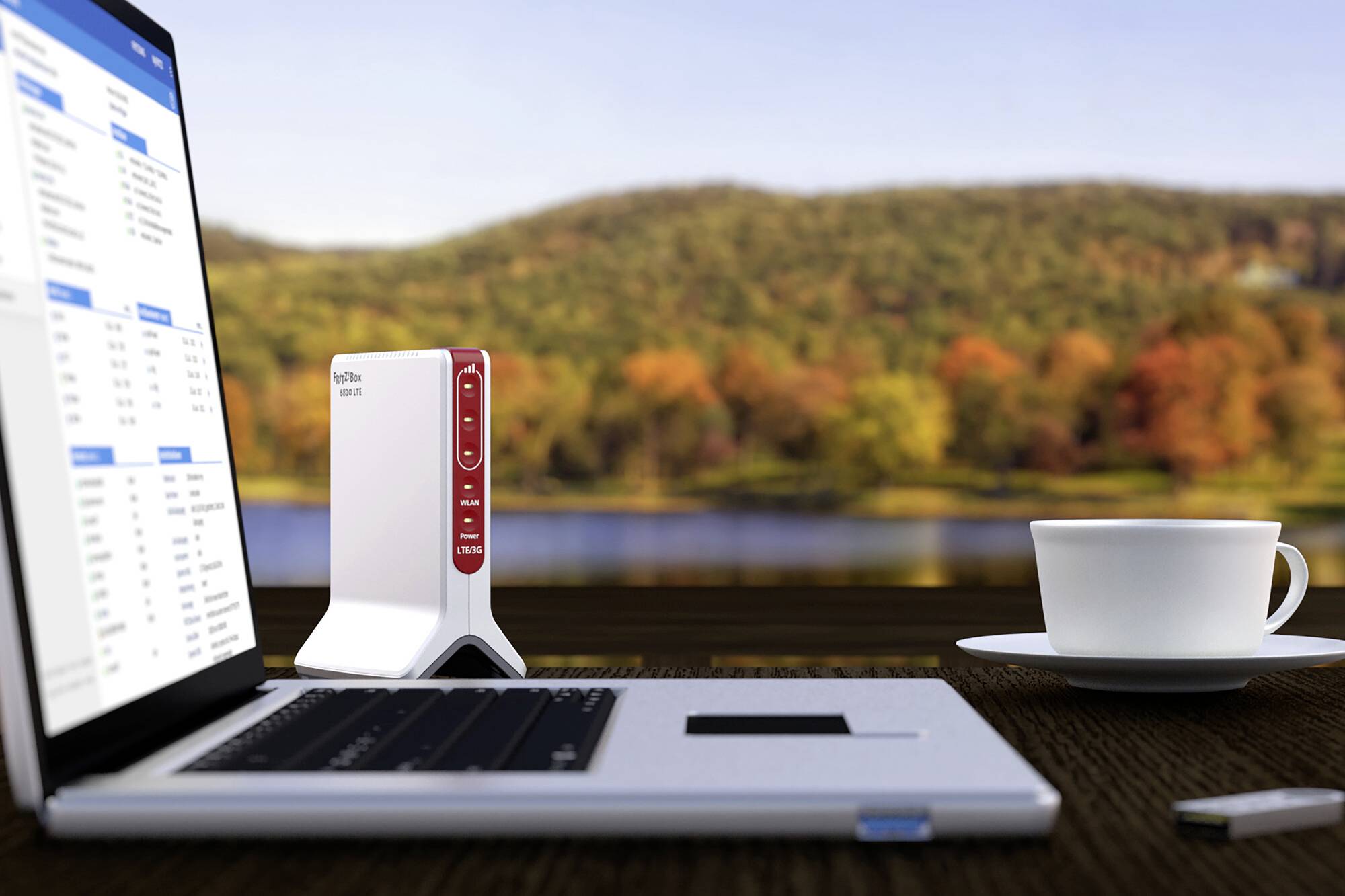 Ein Laptop und ein Router stehen auf einem Tisch mit Blick auf einen See und herbstliche Bäume im Hintergrund. Eine Tasse Kaffee daneben.