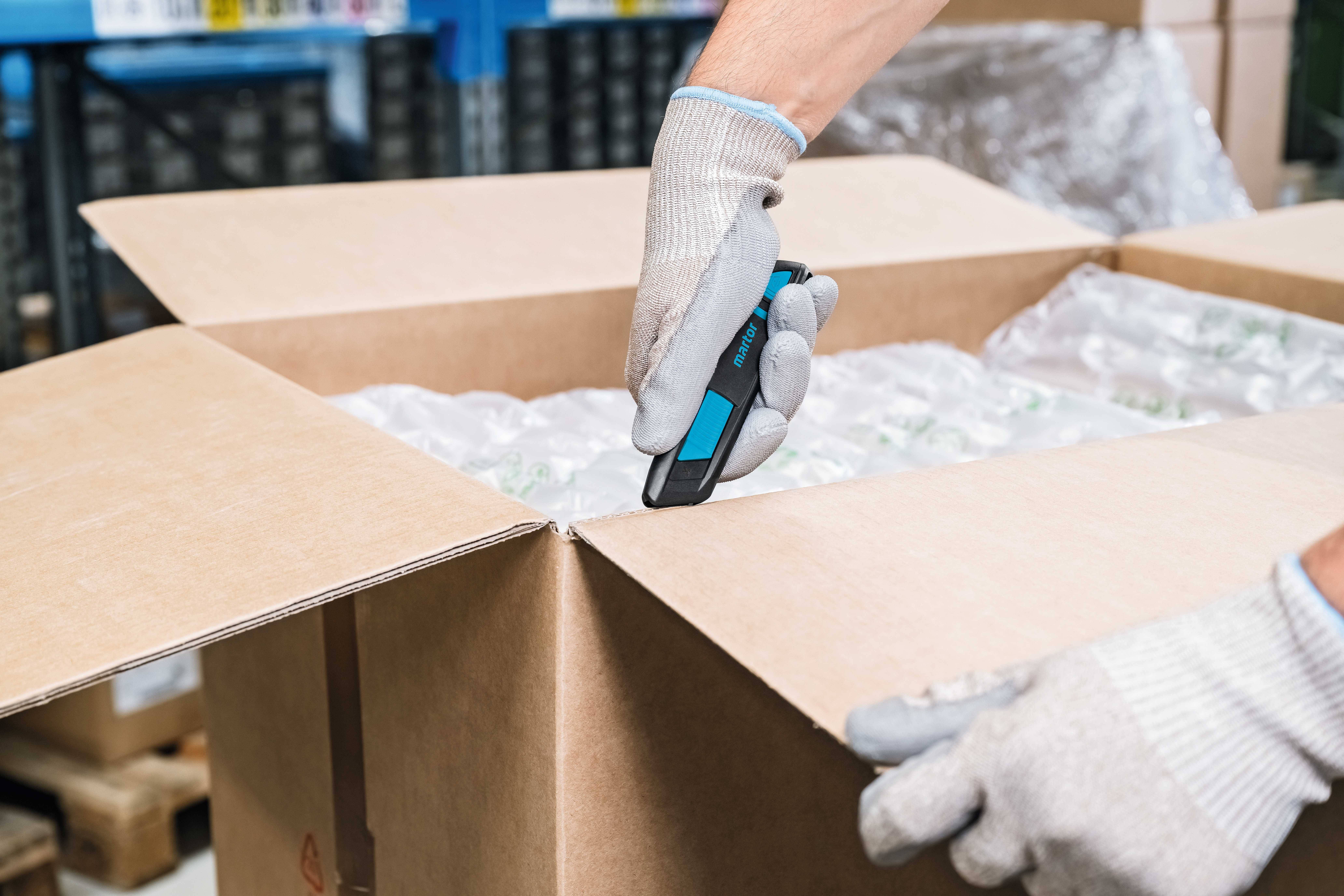 Eine Person öffnet vorsichtig einen großen Karton mit einem blauen Cutter-Messer. Sie trägt dabei graue Arbeitshandschuhe.