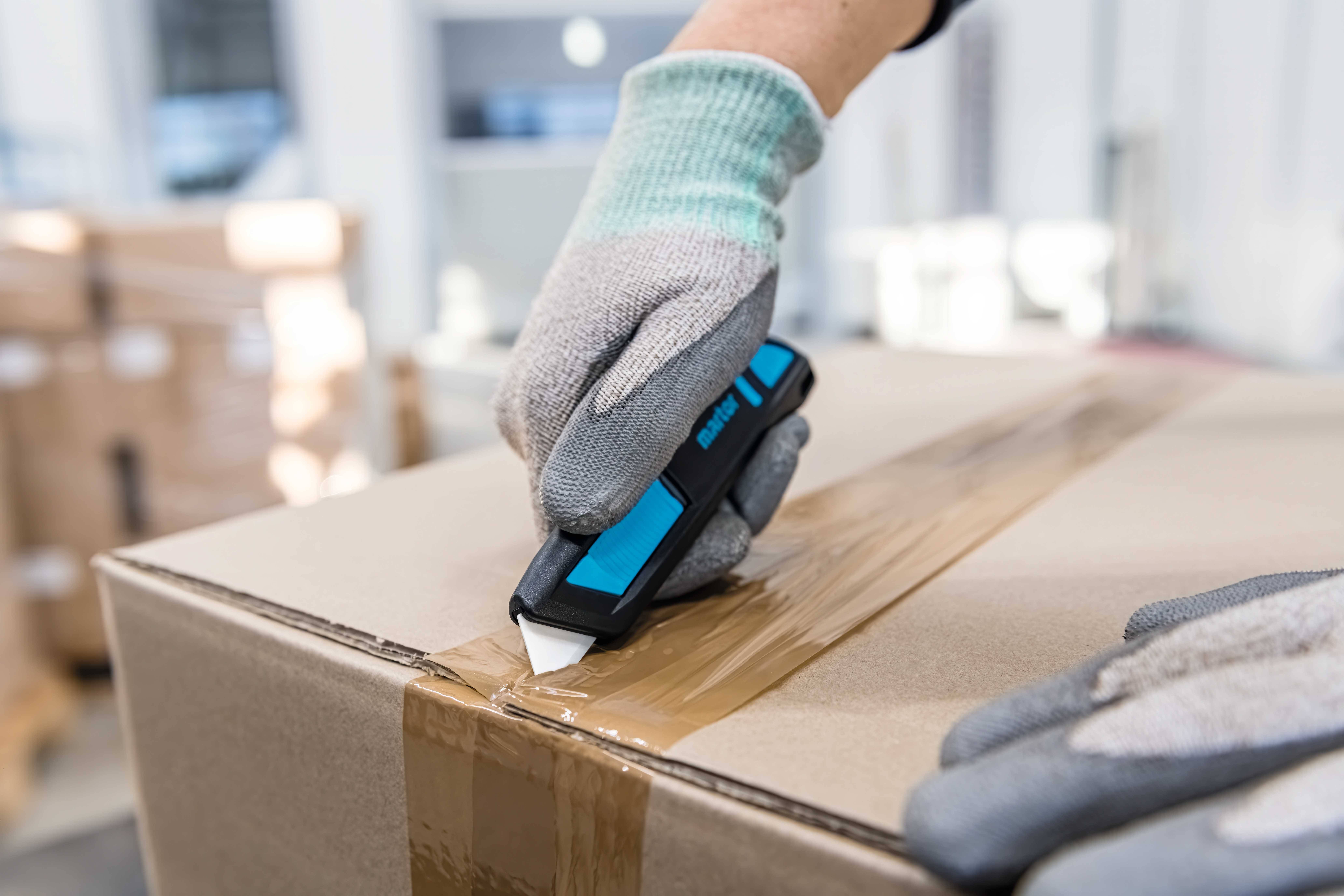 Eine Person in Handschuhen schneidet mit einem Cuttermesser Klebeband auf einem Karton in einem Lager auf.