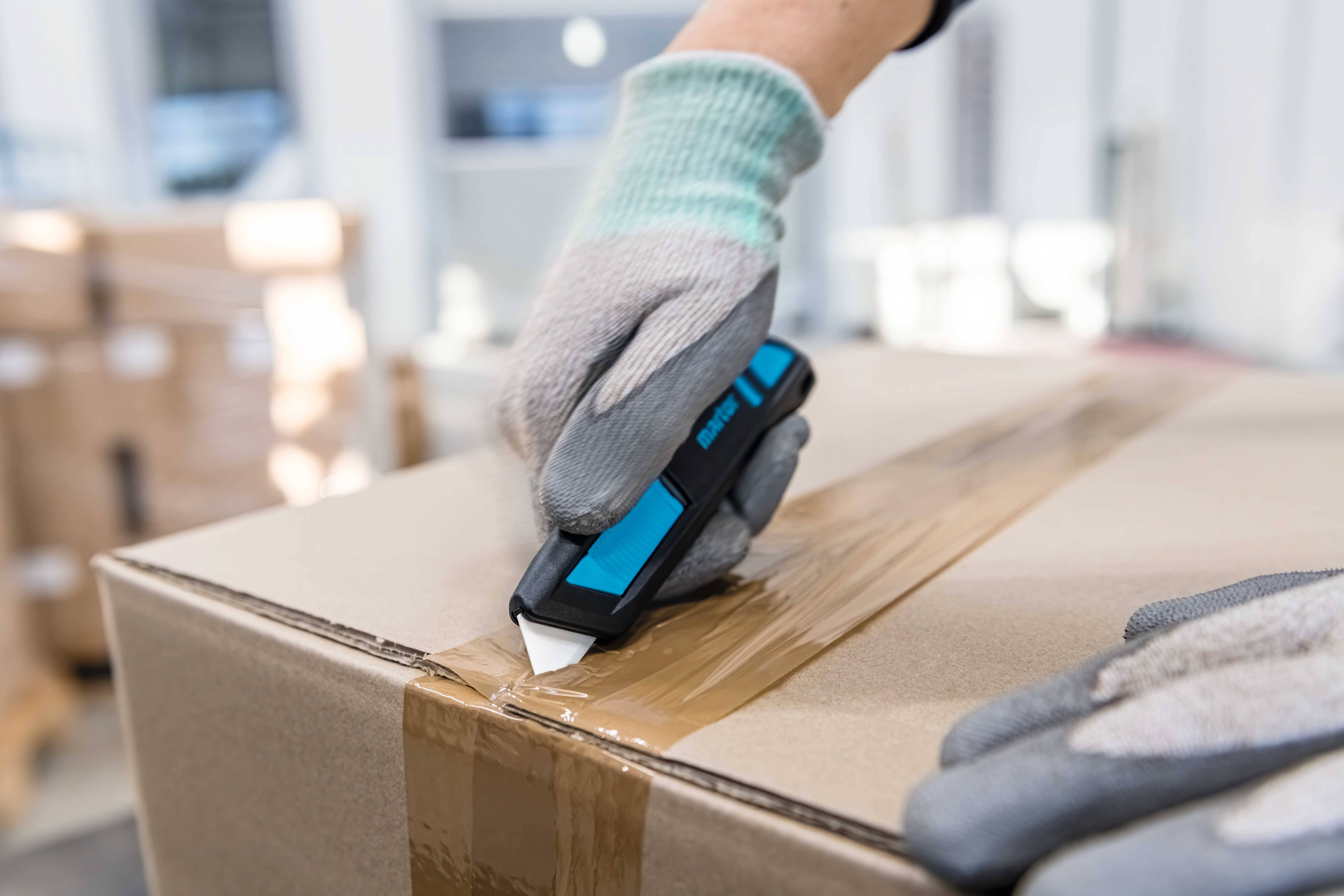 Person in Handschuhen öffnet mit einem blauen Cutter-Messer einen braunen Pappkarton mit Klebeband in einer Lagerumgebung.