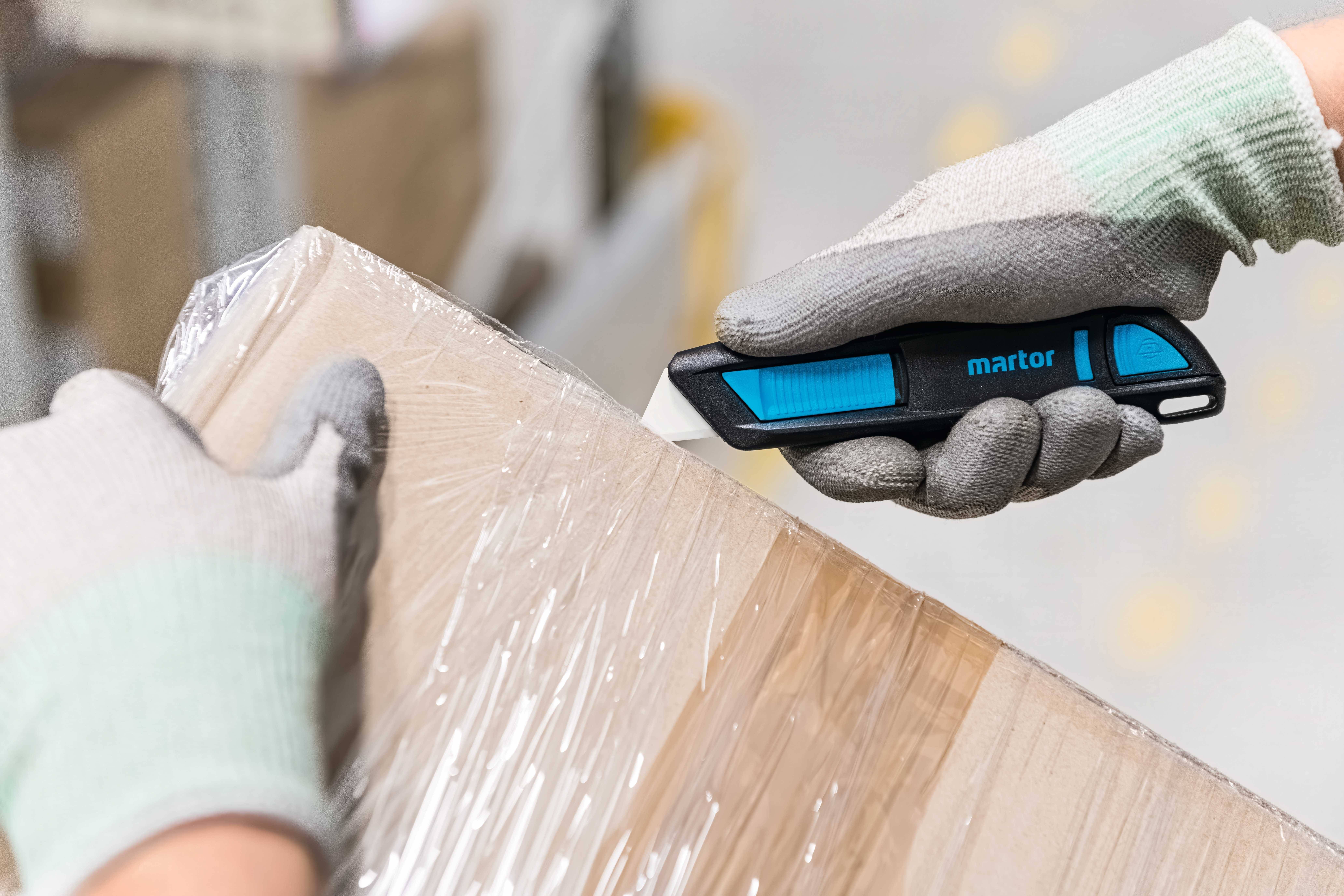 Person mit Handschuhen schneidet Verpackungsfolie eines Kartons mit einem Sicherheitsmesser.