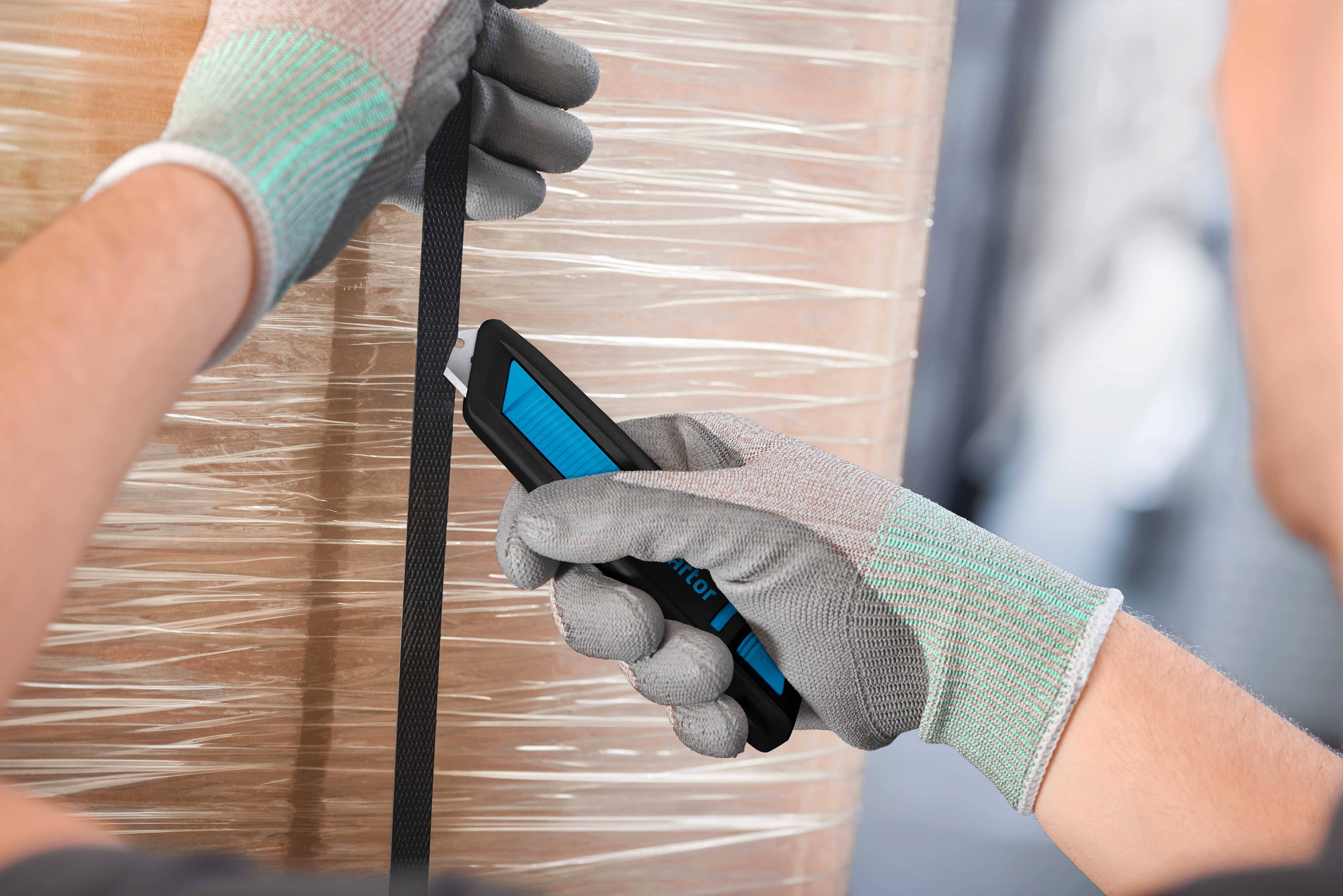 Person mit grauen Handschuhen benutzt ein blau-schwarzes Schneidwerkzeug, um ein schwarzes Band auf einer umwickelten Palette zu schneiden.