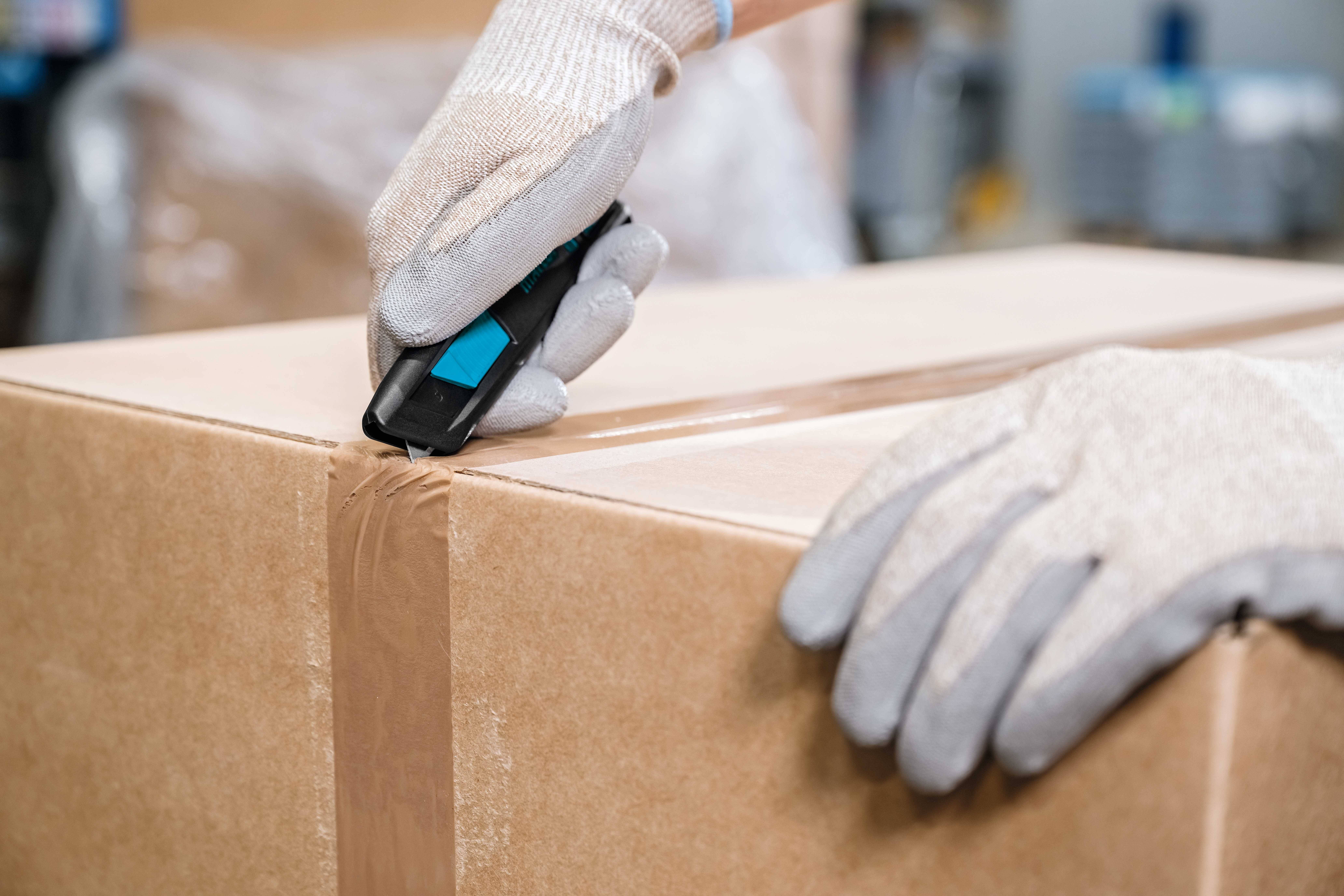 Person mit Handschuhen öffnet Karton mit einem Cutter-Messer.