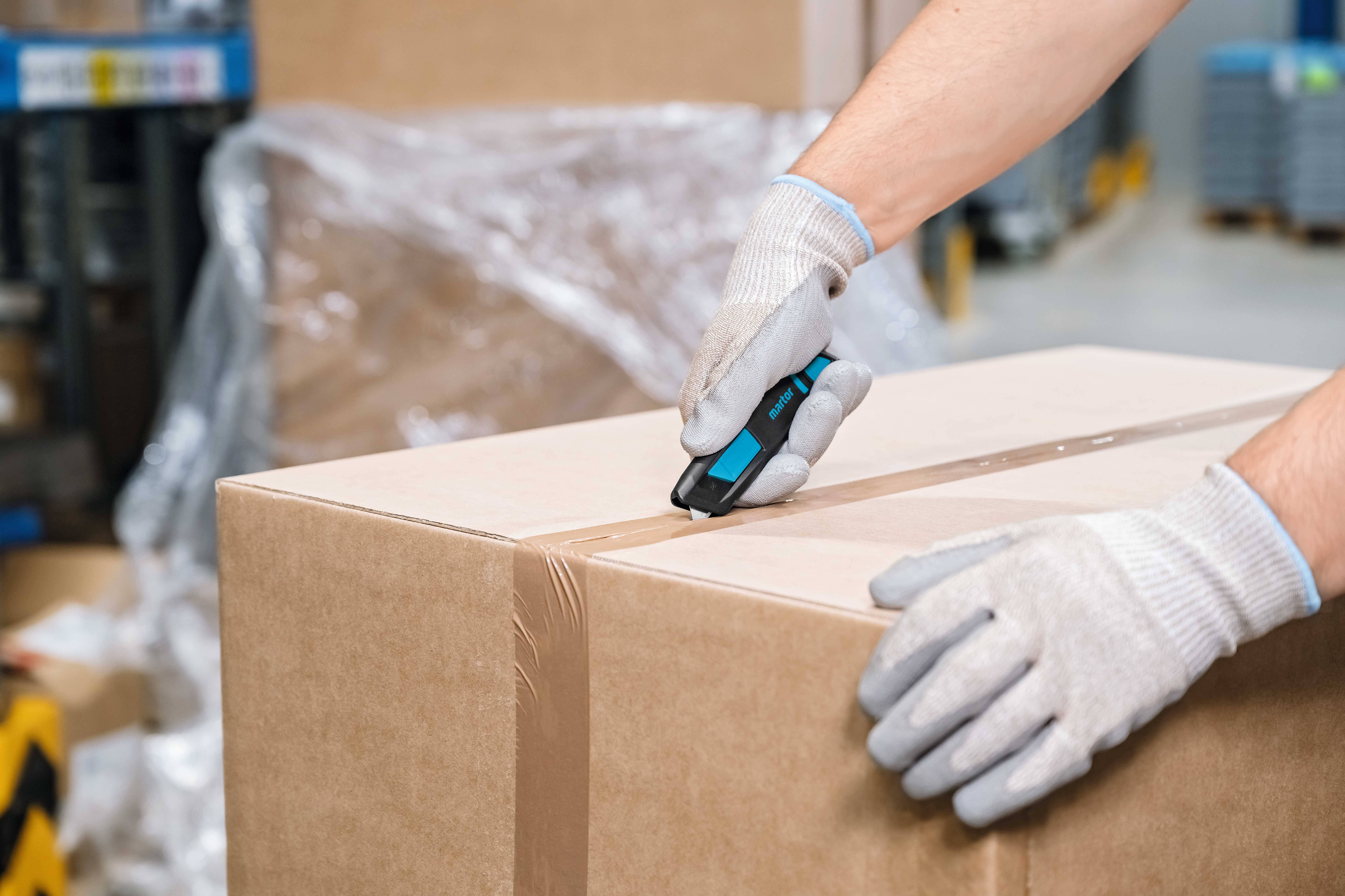 Person in Handschuhen öffnet einen Karton mit einem Messer in einer Lagerumgebung.