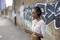 Eine Frau mit Kopfhörern steht vor einer Ziegelwand mit Graffiti und schaut auf ihr Handy. Sie trägt eine weiße Bluse.