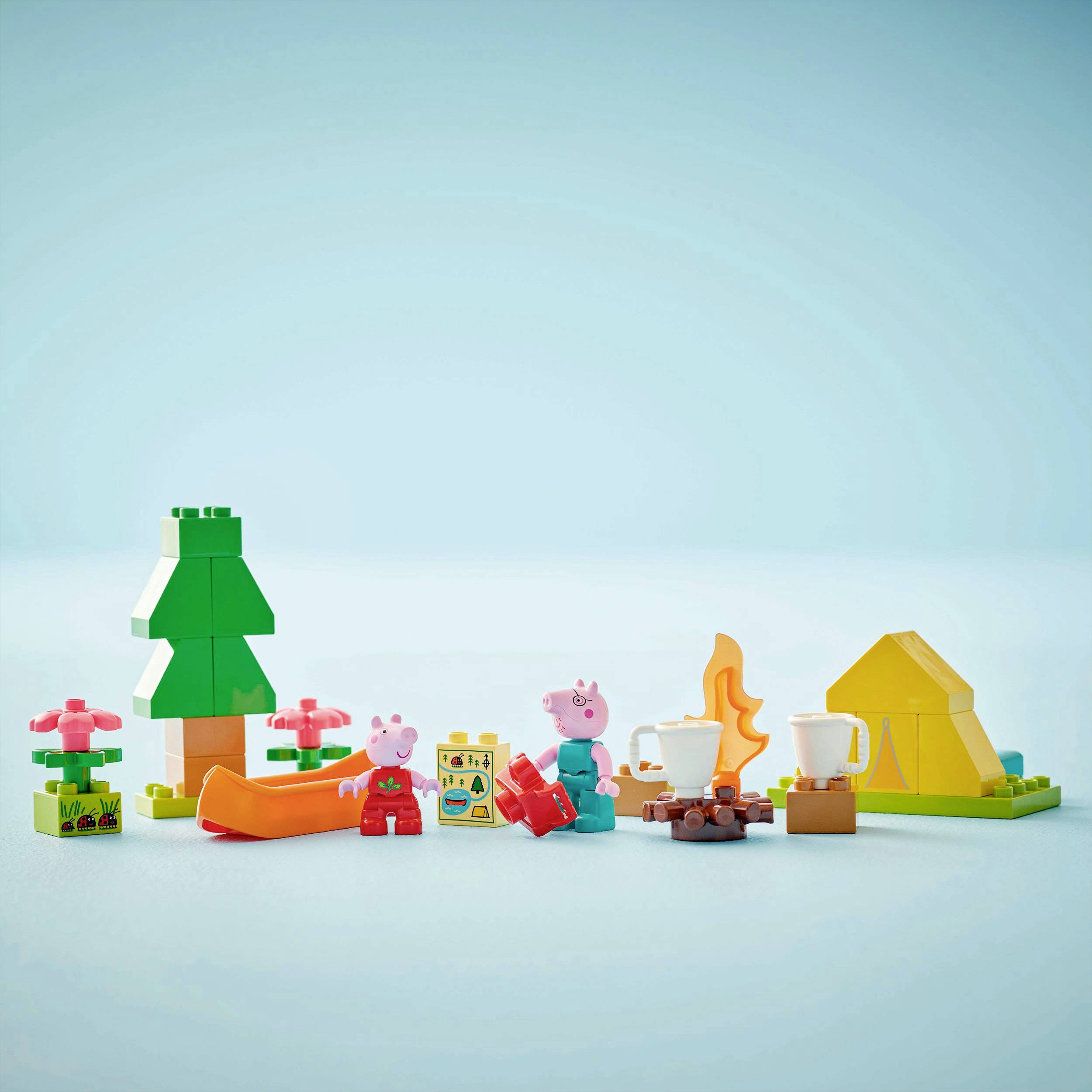 Peppa Wutz und ein anderes Schweinchen sitzen neben einem Zelt am Lagerfeuer aus Bauklötzen, mit einem Baum und Blumen im Hintergrund.