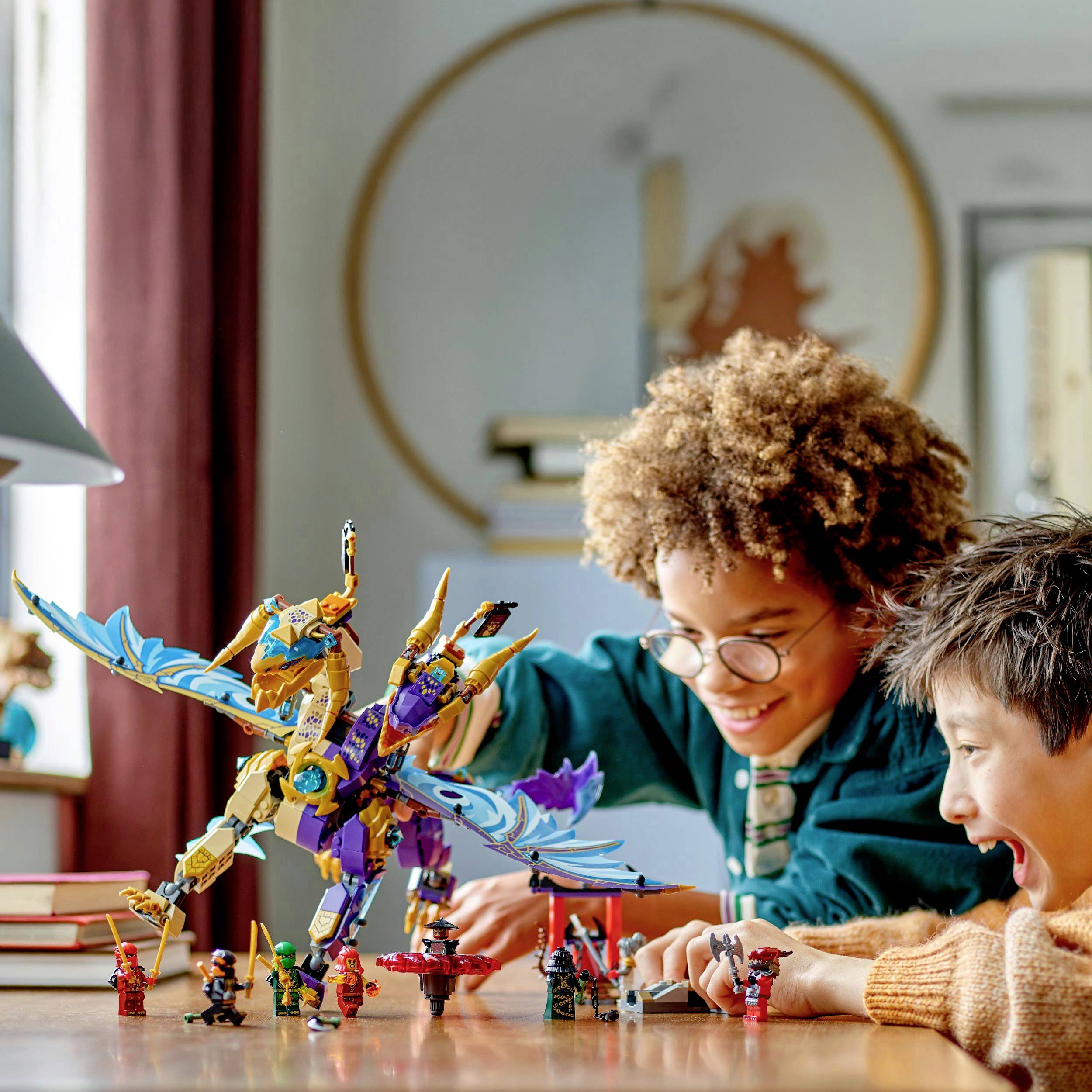 Ein Kind mit lockigem Haar und Brille und ein weiteres Kind spielen lachend mit bunten Robotermodellen auf einem Tisch in einem Zimmer.