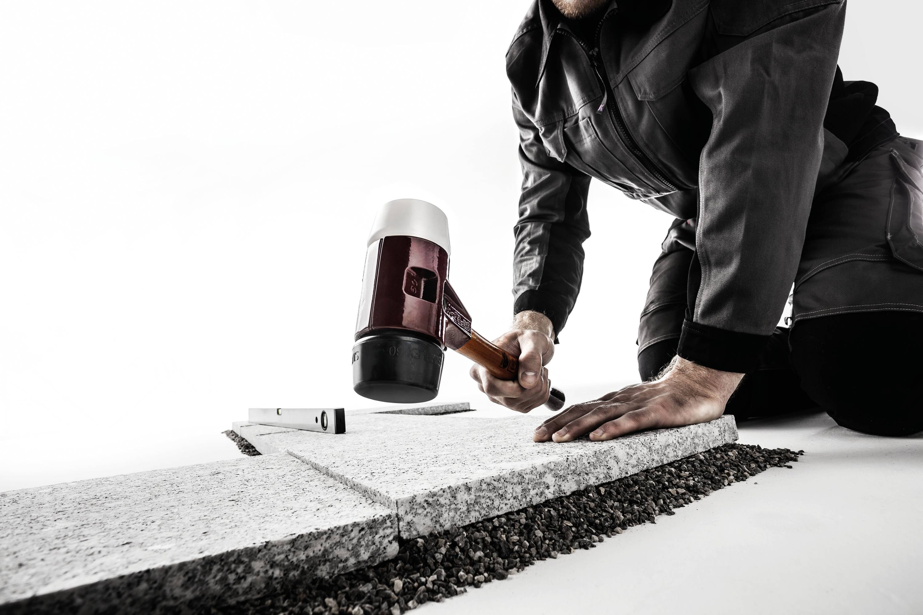 Eine Person kniet auf dem Boden und benutzt einen Hammer, um Pflastersteine auf einem Kiesbett zu verlegen.