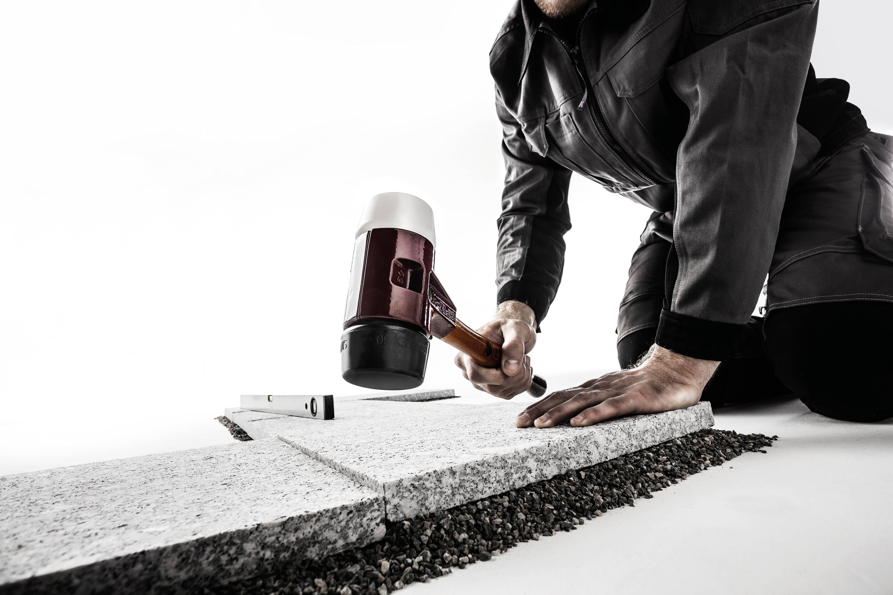 Eine Person kniet und bearbeitet mit einem Hammer einen Steinbodenbelag, um ihn anzupassen. Der Hintergrund ist weiß.