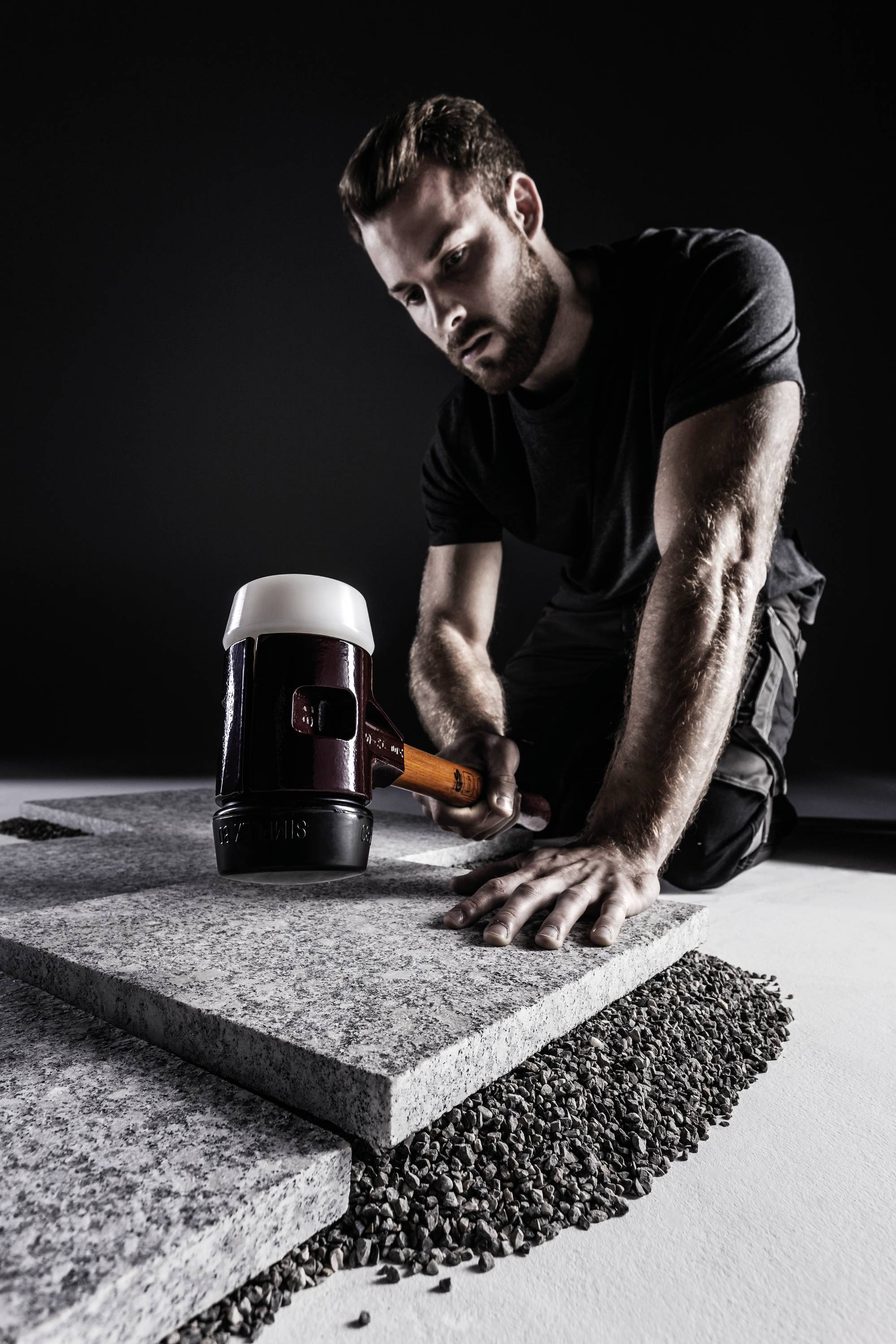 Ein Mann kniet auf dem Boden und benutzt einen Hammer, um Fliesen auf Schotter zu verlegen. Die Szene ist in einem dunklen Raum.