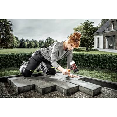 Eine Frau kniet auf einer Terrasse und verlegt dunkelgraue Pflastersteine mit einem Gummihammer. Im Hintergrund sind ein Garten und ein modernes Haus zu sehen.