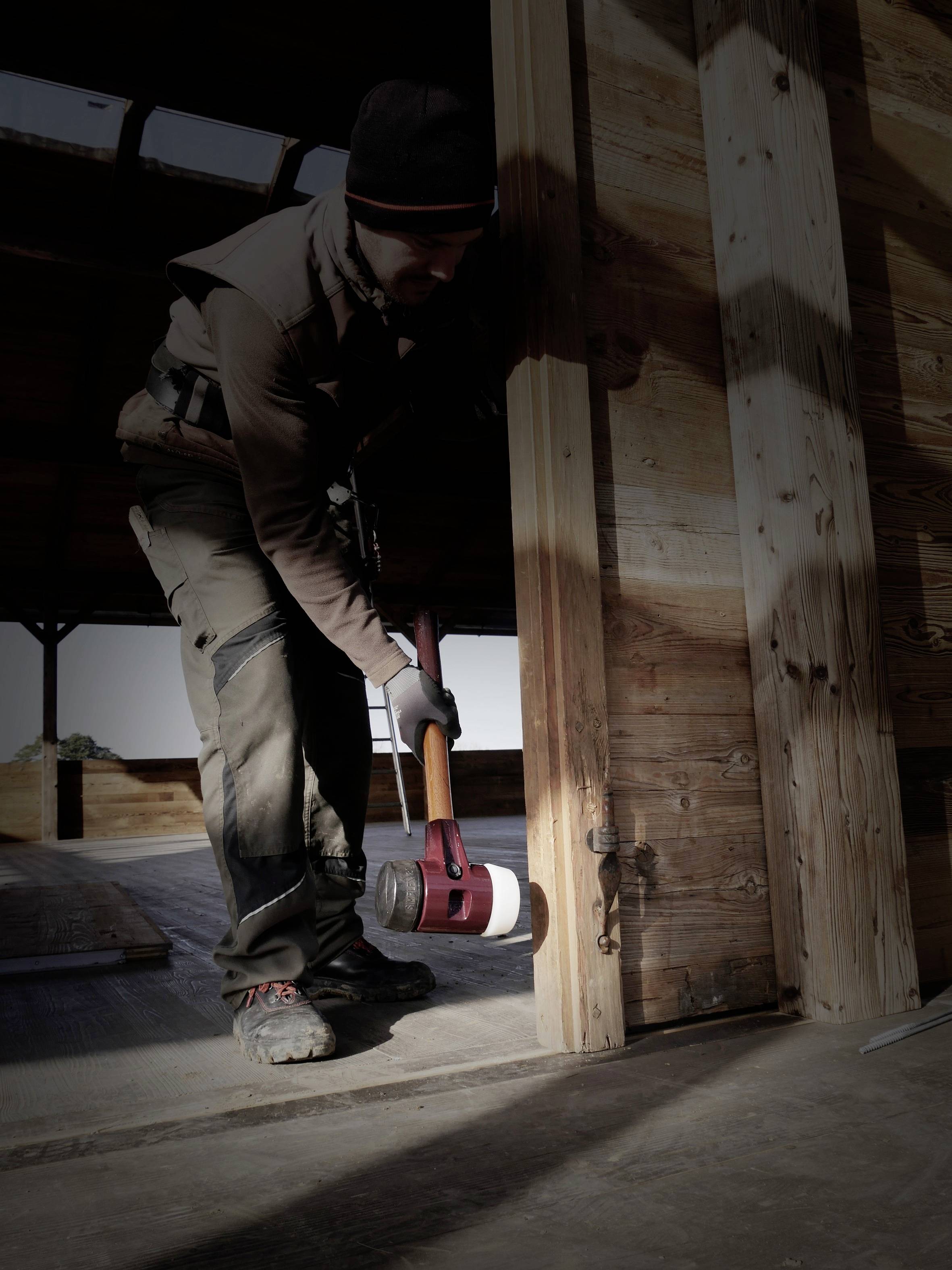 Eine Person, die in einem halbfertigen Holzgebäude mit einem Hammer auf den Rahmen klopft, wahrscheinlich um es zu errichten oder zu reparieren.