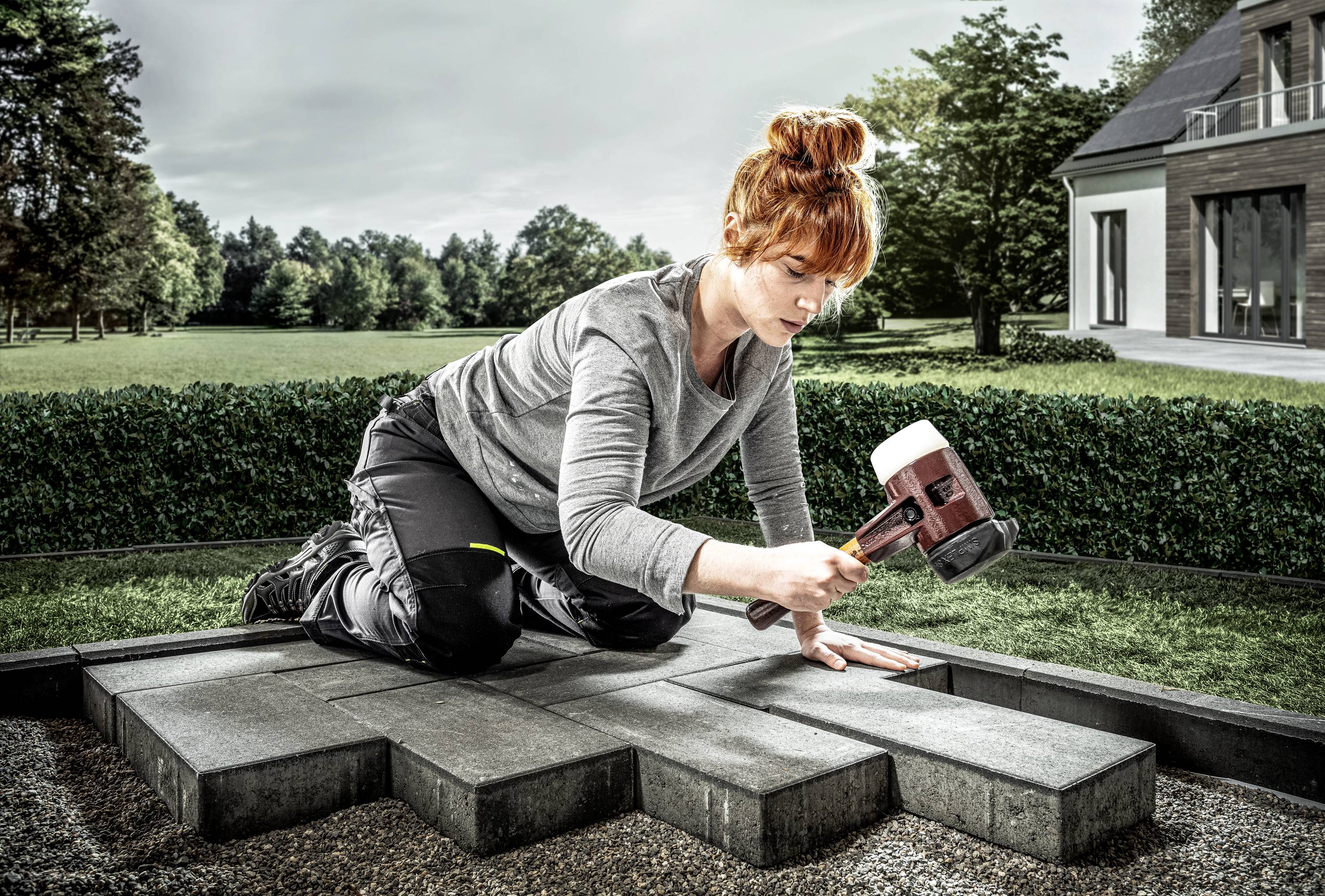 Frau kniet im Garten und pflastert Steine mit Gummihammer. Im Hintergrund ein modernes Haus und Bäume.