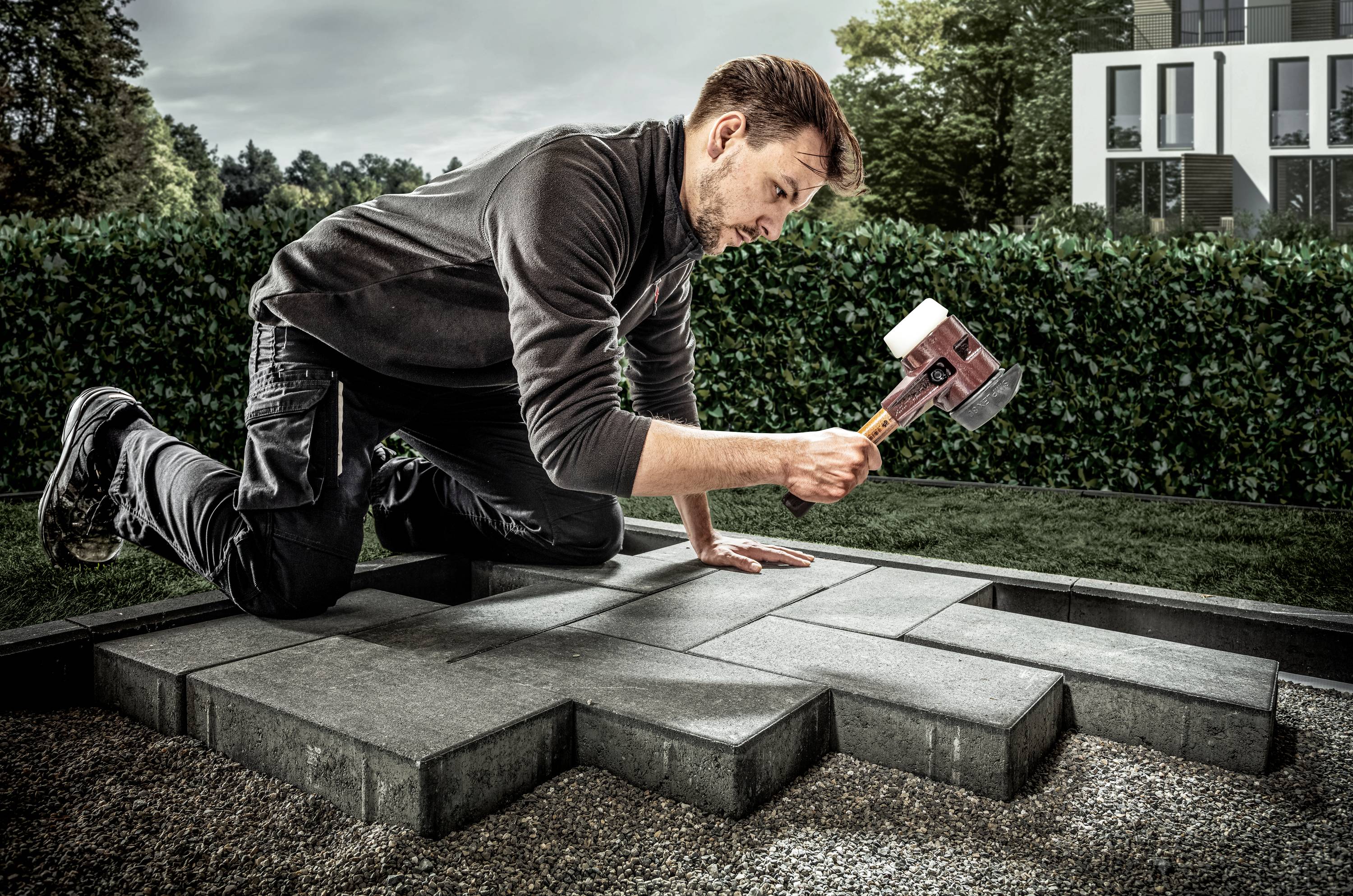Ein Mann kniet auf dem Boden und benutzt einen großen Hammer, um Pflastersteine im Garten zu verlegen. Im Hintergrund sind Büsche und ein modernes Haus sichtbar.