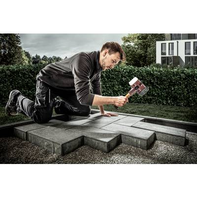Ein Mann kniet auf dem Boden und benutzt einen großen Hammer, um Pflastersteine im Garten zu verlegen. Im Hintergrund sind Büsche und ein modernes Haus sichtbar.