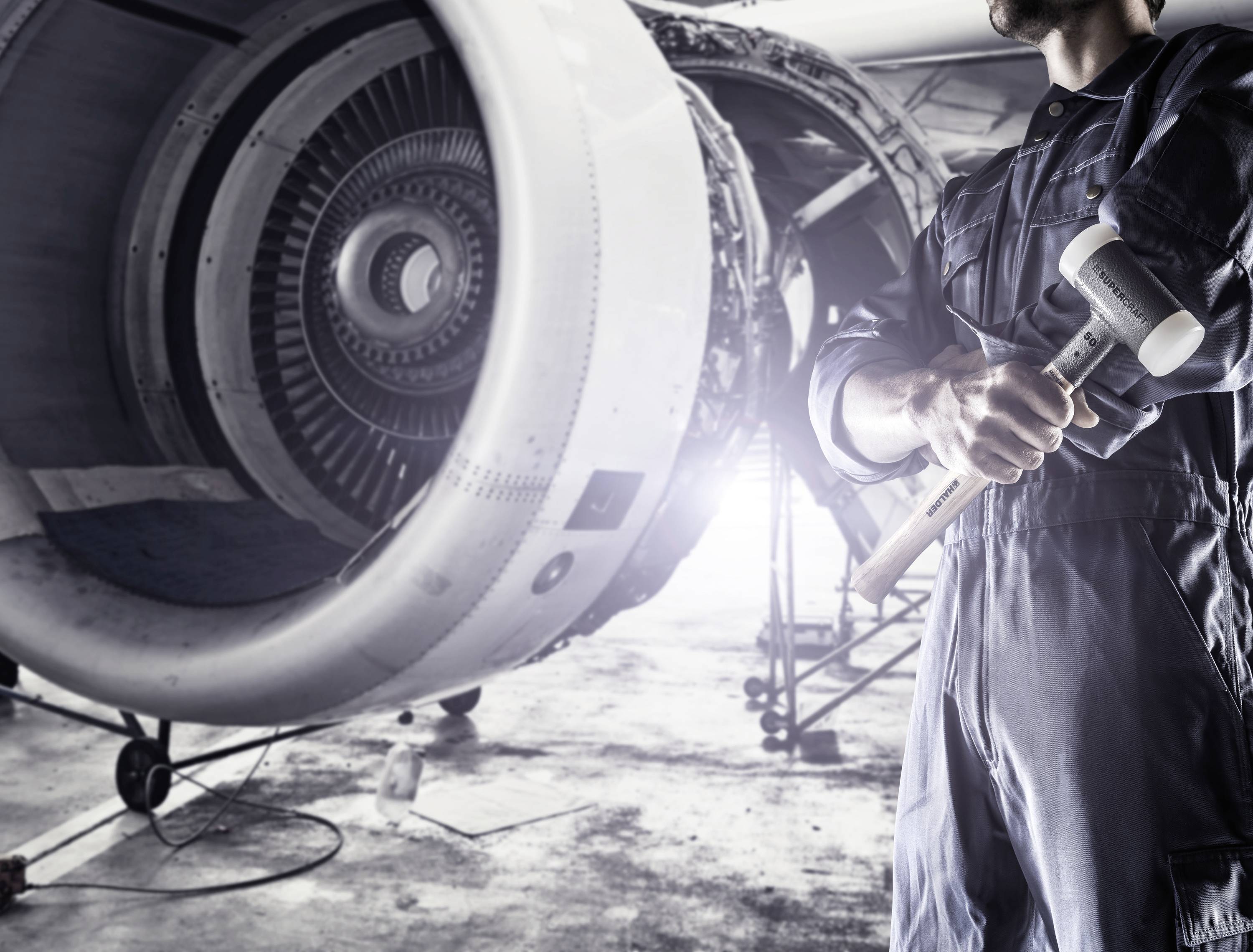 Ein Techniker in Arbeitskleidung hält einen Hammer und steht vor einem Flugzeugtriebwerk in einer Werkstattumgebung.