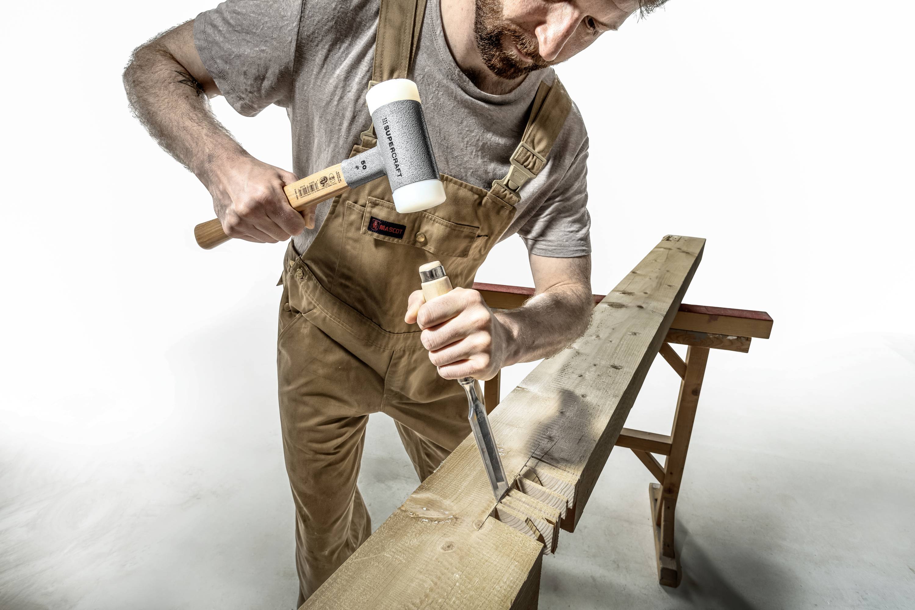 Ein Handwerker in Arbeitskleidung benutzt einen Holzhammer und Meißel, um präzise Schnitte in ein Holzbrett auf einer Werkbank zu machen.