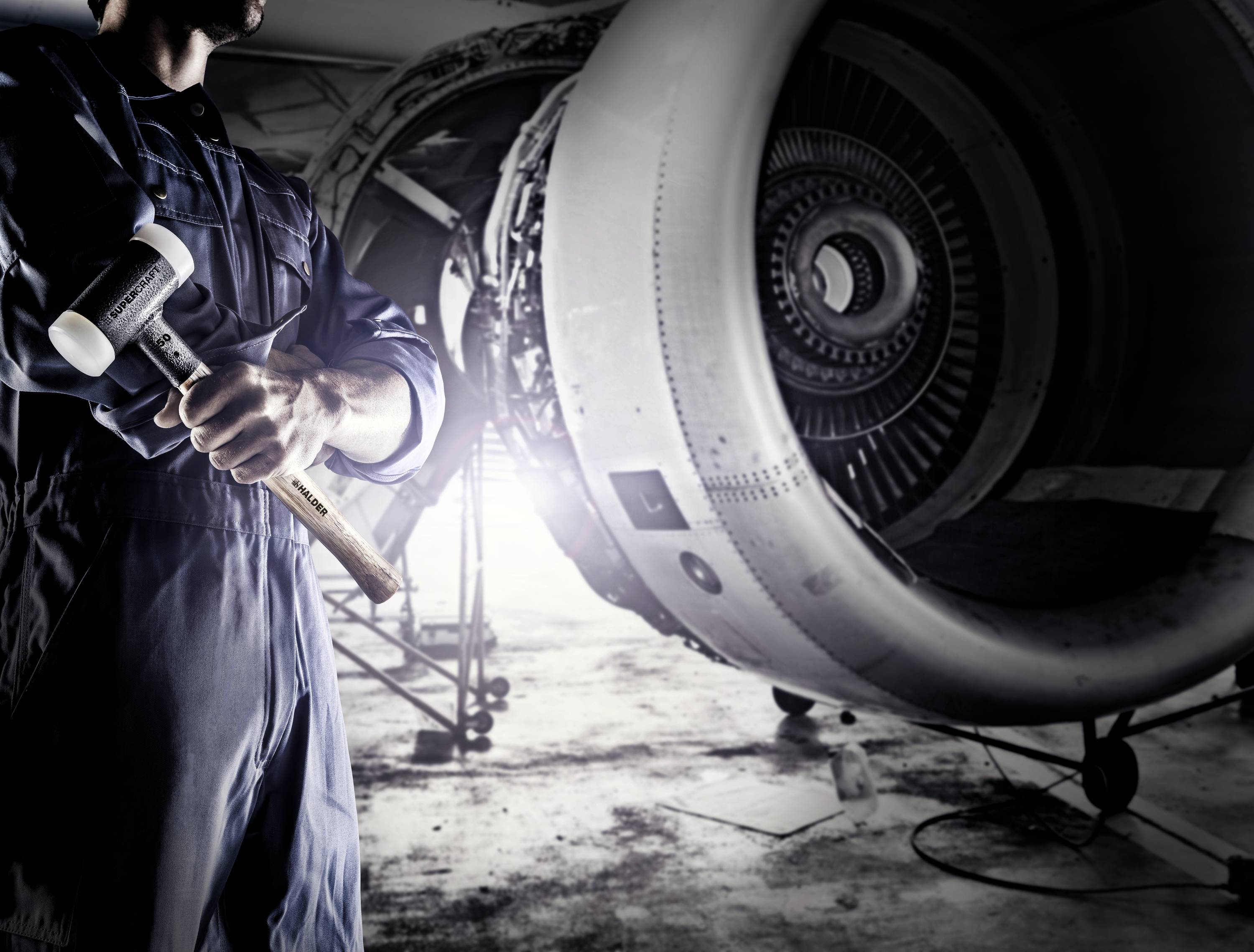 Eine Person im blauen Overall hält einen Hammer vor einem großen Flugzeugtriebwerk. Die Umgebung wirkt industriell und technisch.
