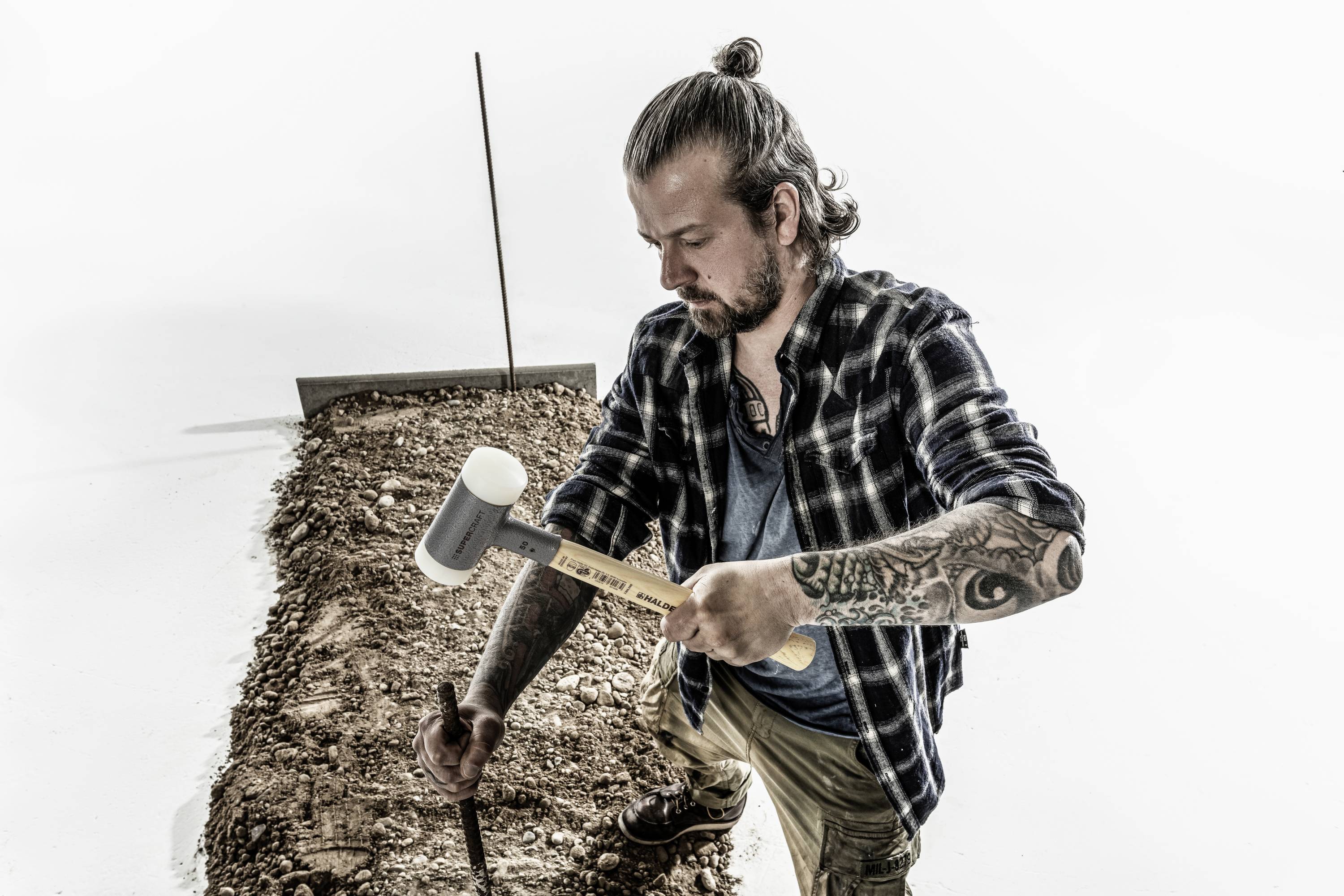 Ein Mann mit Tattoos hämmert mit einem Gummihammer einen Pflock in den Boden. Er trägt ein kariertes Hemd und hat einen Dutt.