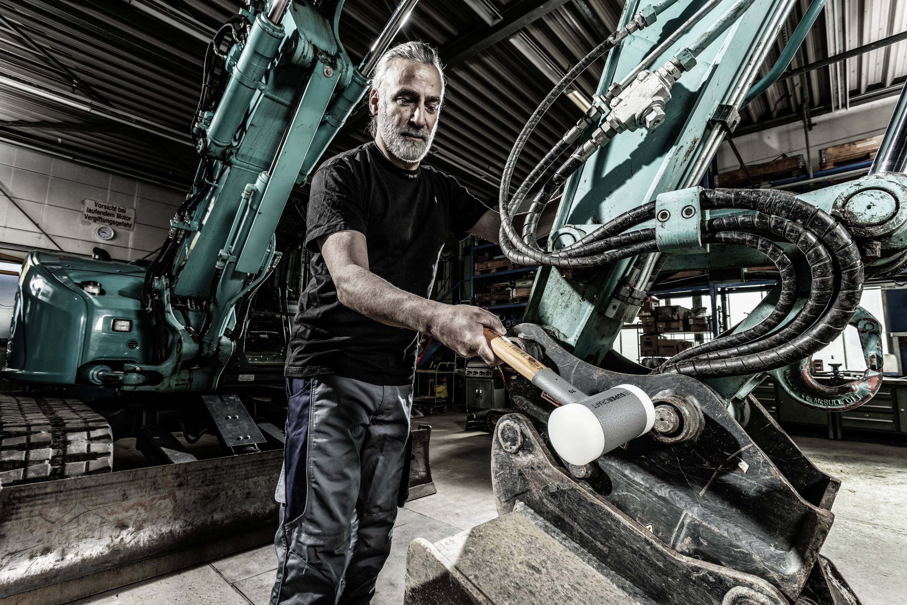 Ein Mann arbeitet in einer Werkstatt an einem großen Bagger, hält einen großen Hammer. Die Umgebung wirkt industriell.