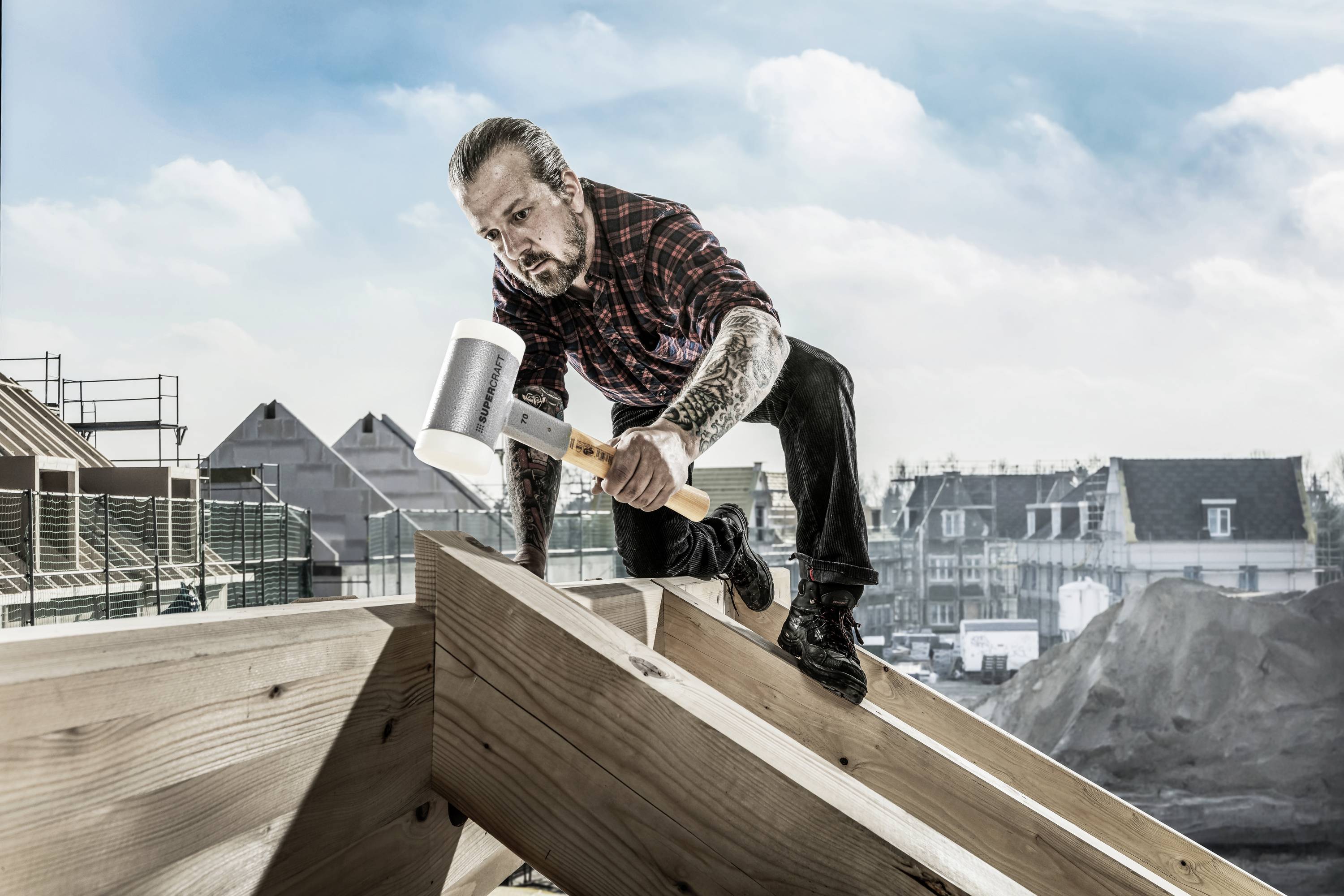 Ein Mann auf dem Dach eines Gebäudes arbeitet mit Hammer und Klammerzange. Im Hintergrund sind Gebäude und ein klarer Himmel sichtbar.