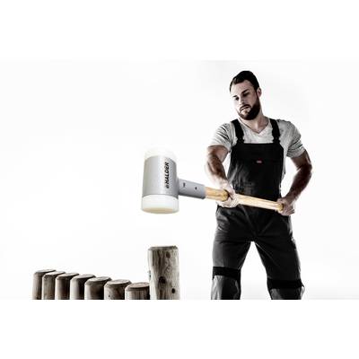 Ein Mann in Arbeitskleidung benutzt einen großen Hammer, um Holzpfähle zu bearbeiten. Die Szene zeigt handwerkliche Arbeit.