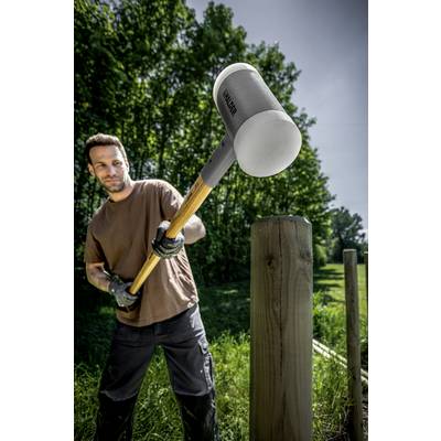 Ein Mann schlägt mit einem großen Hammer auf einen Holzpfosten im Freien ein. Im Hintergrund sind Bäume und Gras zu sehen.