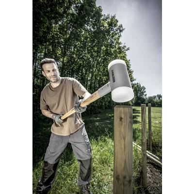 Mann in Arbeitskleidung schlägt mit einem großen Hammer einen Holzpfosten in den Boden, um einen Zaun in einer grünen Umgebung zu errichten.